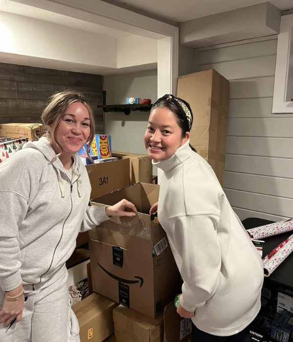 Two women standing among boxes, one wearing a light gray hoodie and the other a white sweatshirt, in a room with wrapping paper and shelves.