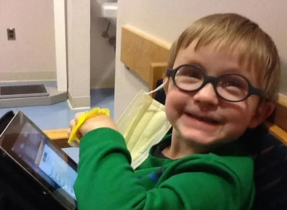 A young boy with glasses smiling and lying in a hospital bed, holding a tablet in one hand.