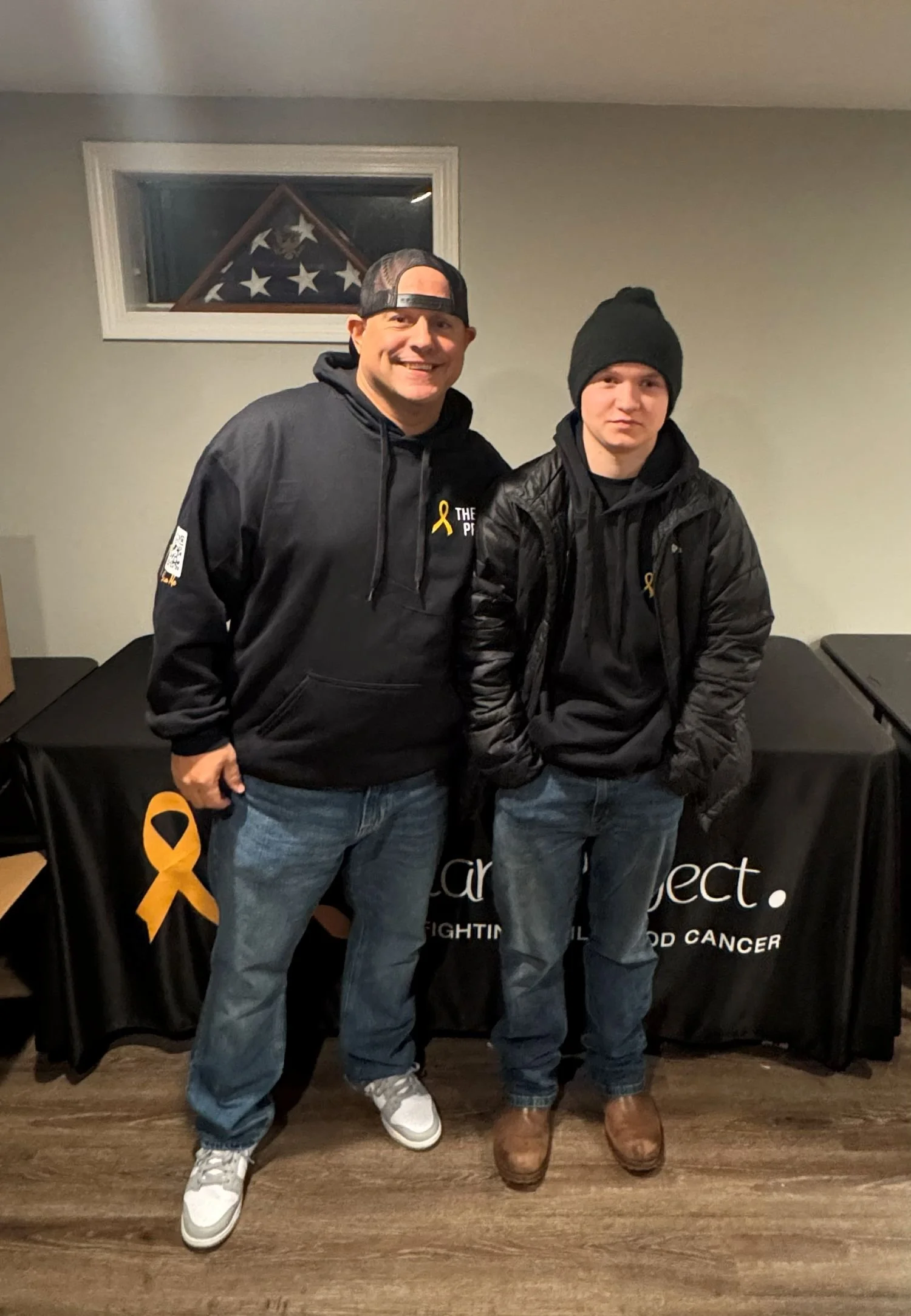 Two men standing side by side in front of a table with a black tablecloth featuring yellow ribbons and white text. They are wearing black hoodies with yellow ribbons on them. The man on the left is smiling and wearing a black cap backwards, while the man on the right is wearing a black beanie and has his hands in his pockets. There is a small window in the background with a flag folded into a triangular shape.