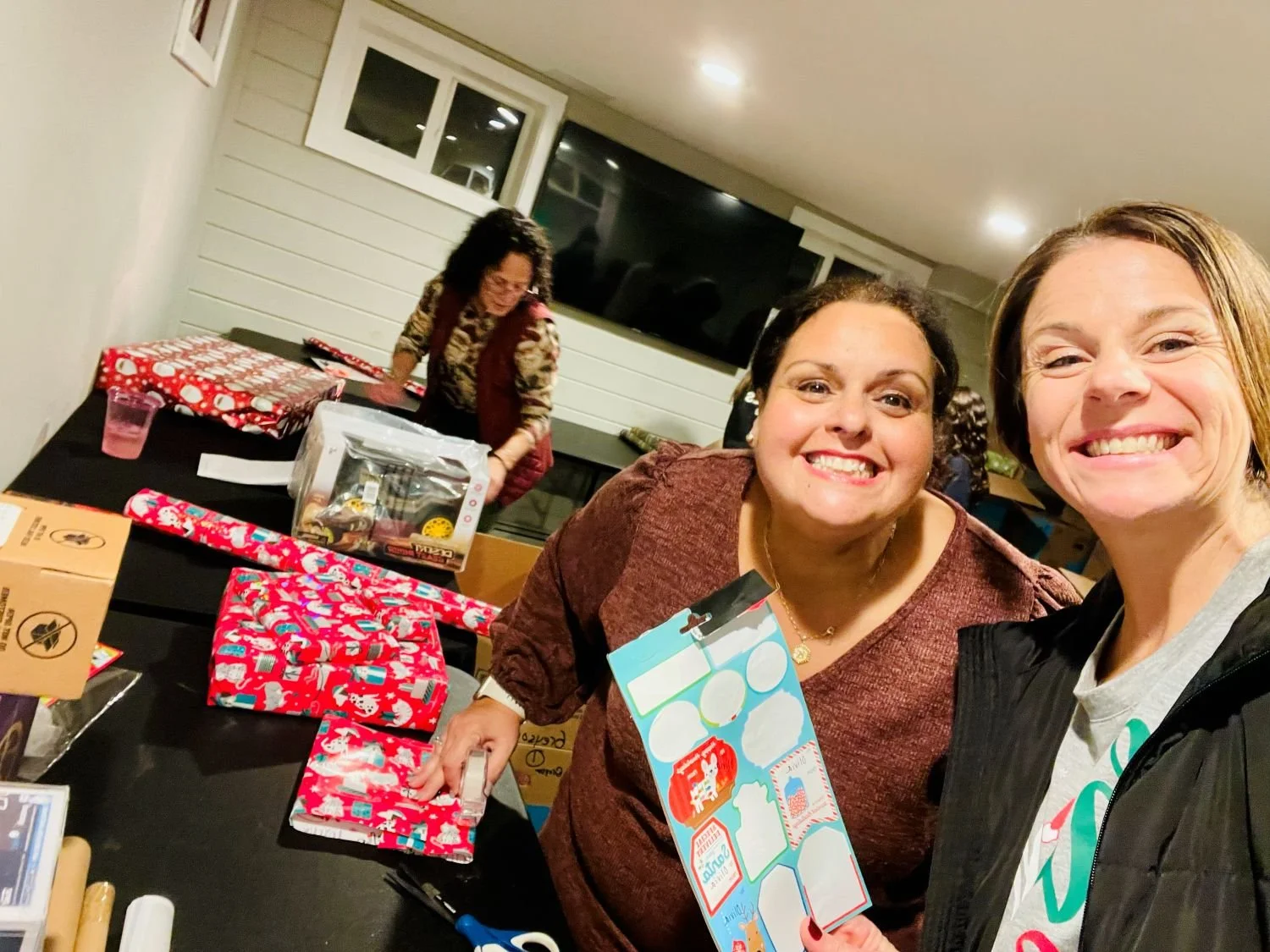 Two women smiling and taking a selfie at a Christmas gift exchange, with wrapped presents on the table and a woman in the background preparing gifts.