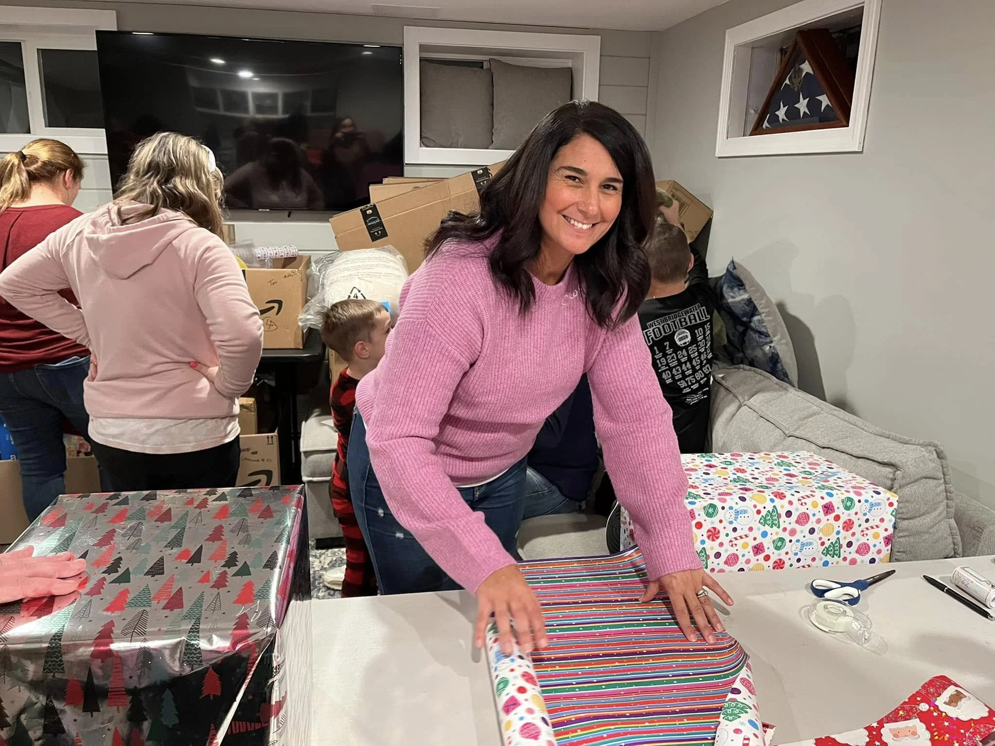 A woman smiling, wrapping presents at a holiday gathering, with wrapped gifts on the table and other people in the background preparing for Christmas.