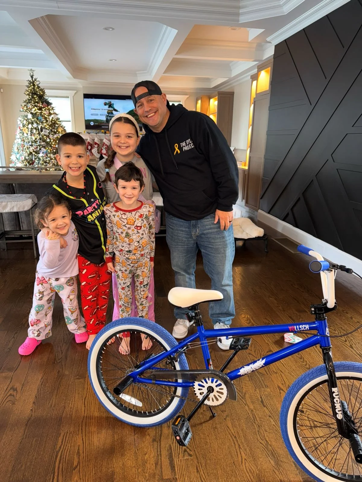 Man and five children in pajamas posing together inside a home with a Christmas tree in the background, Christmas stockings, and a blue bicycle in the foreground.