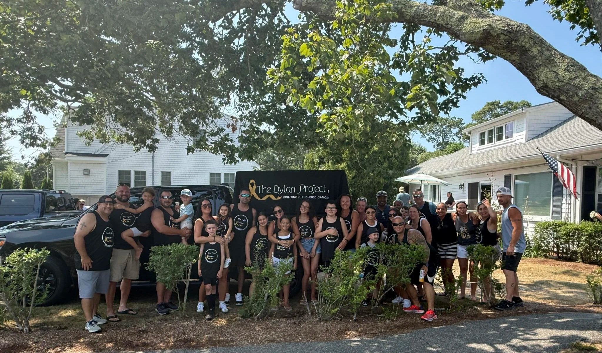 Group of people gathered outdoors in front of a black van, with a sign that reads 'The Dylan Project fighting childhood cancer,' under a large tree, in a residential neighborhood.