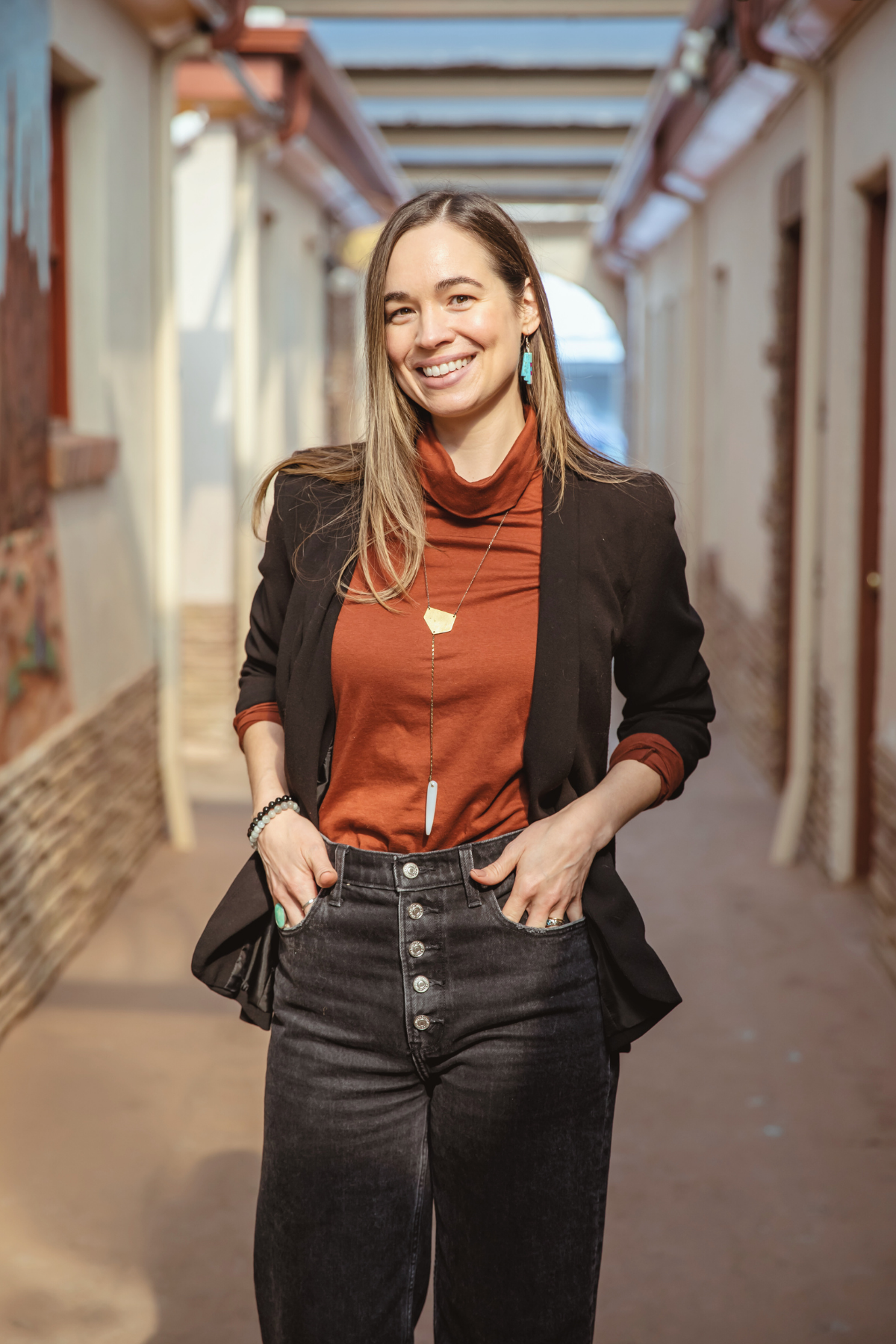 A smiling woman with long light brown hair, wearing a black blazer, rust-colored turtleneck, and high-waisted black jeans, standing in an outdoor alleyway with a wooden roof structure.