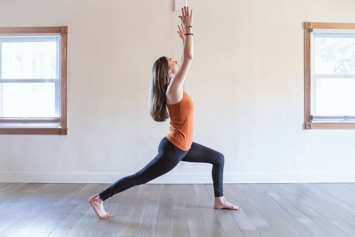 Woman practicing yoga indoors, holding a pose with arms raised and one leg stretched forward.