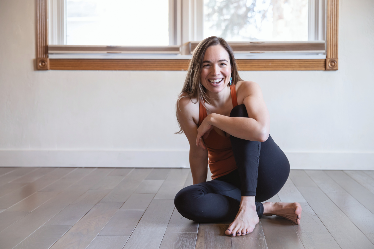 A woman in workout clothes sitting on the wooden floor, smiling at the camera, with a window behind her.