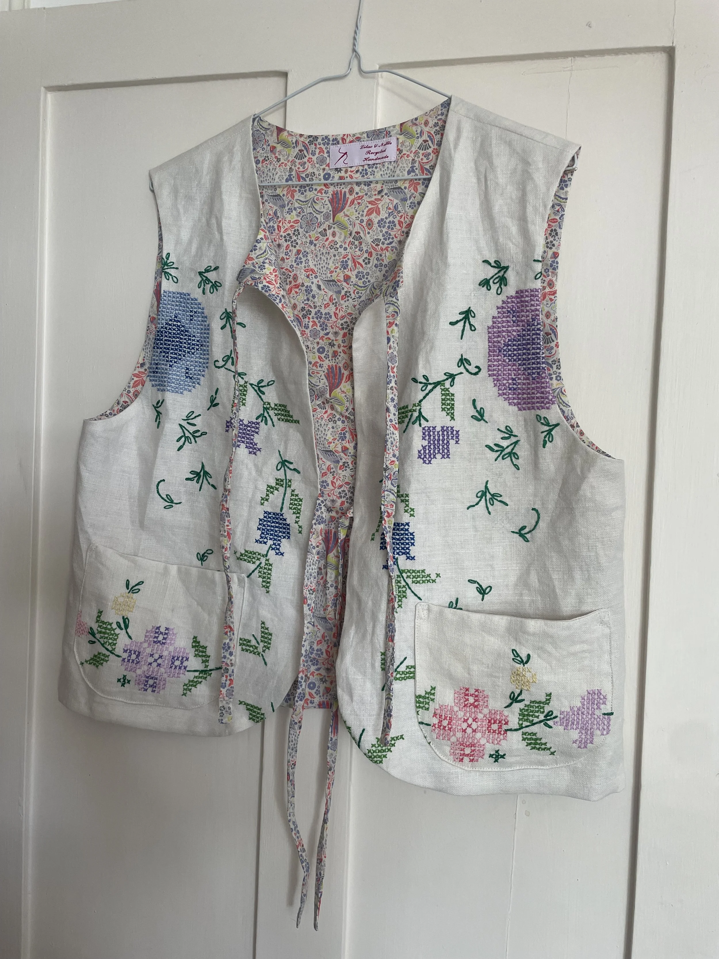 A sleeveless, embroidered, white linen vest hanging on a hanger against a white door. The vest features colorful floral embroidery with purple, pink, blue, yellow, and green threads, and has large front pockets.