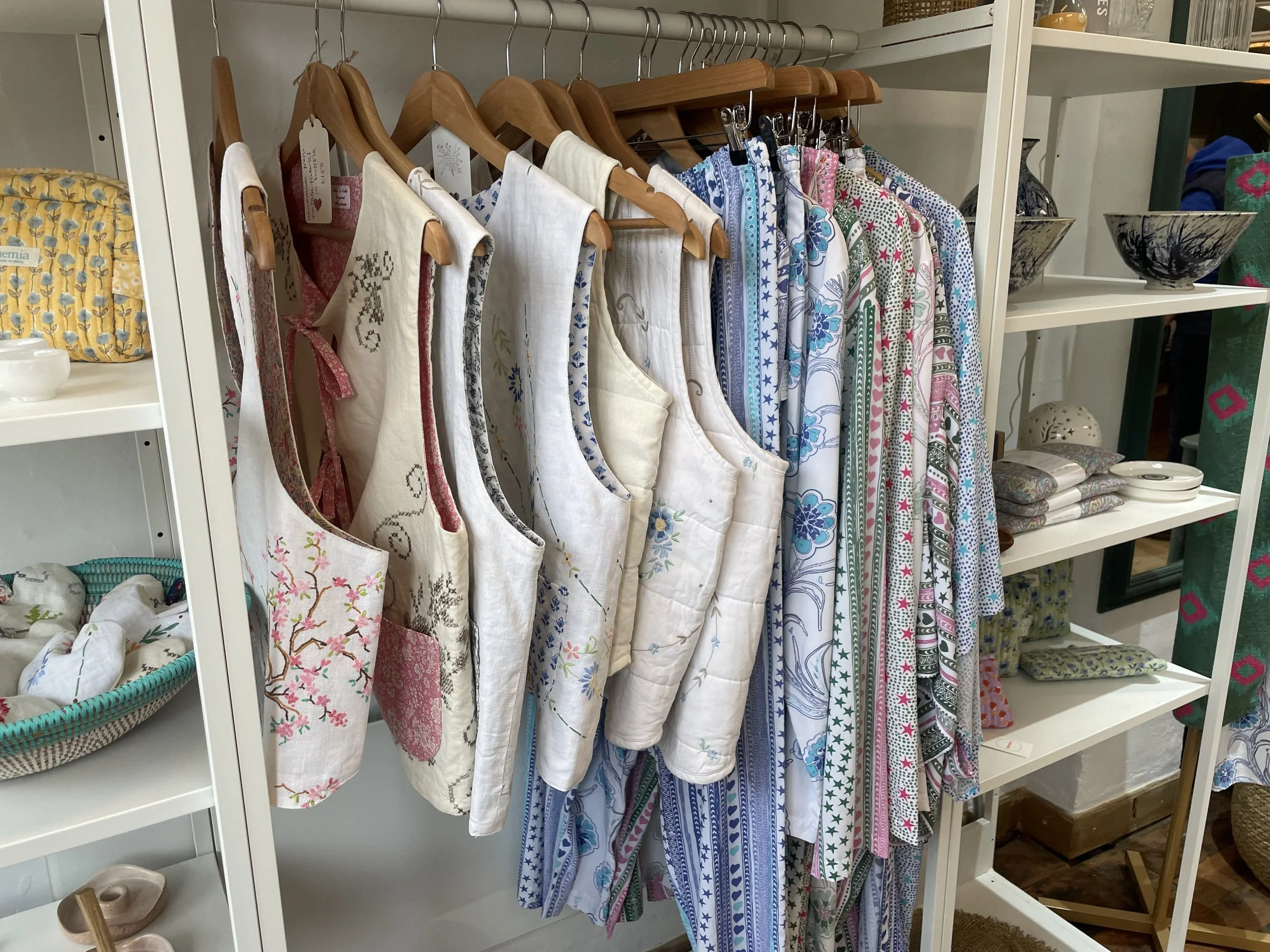 A retail display of various embroidered and printed fabric aprons and shirts hanging on wooden hangers and hooks, with shelves of bowls and folded textiles nearby.