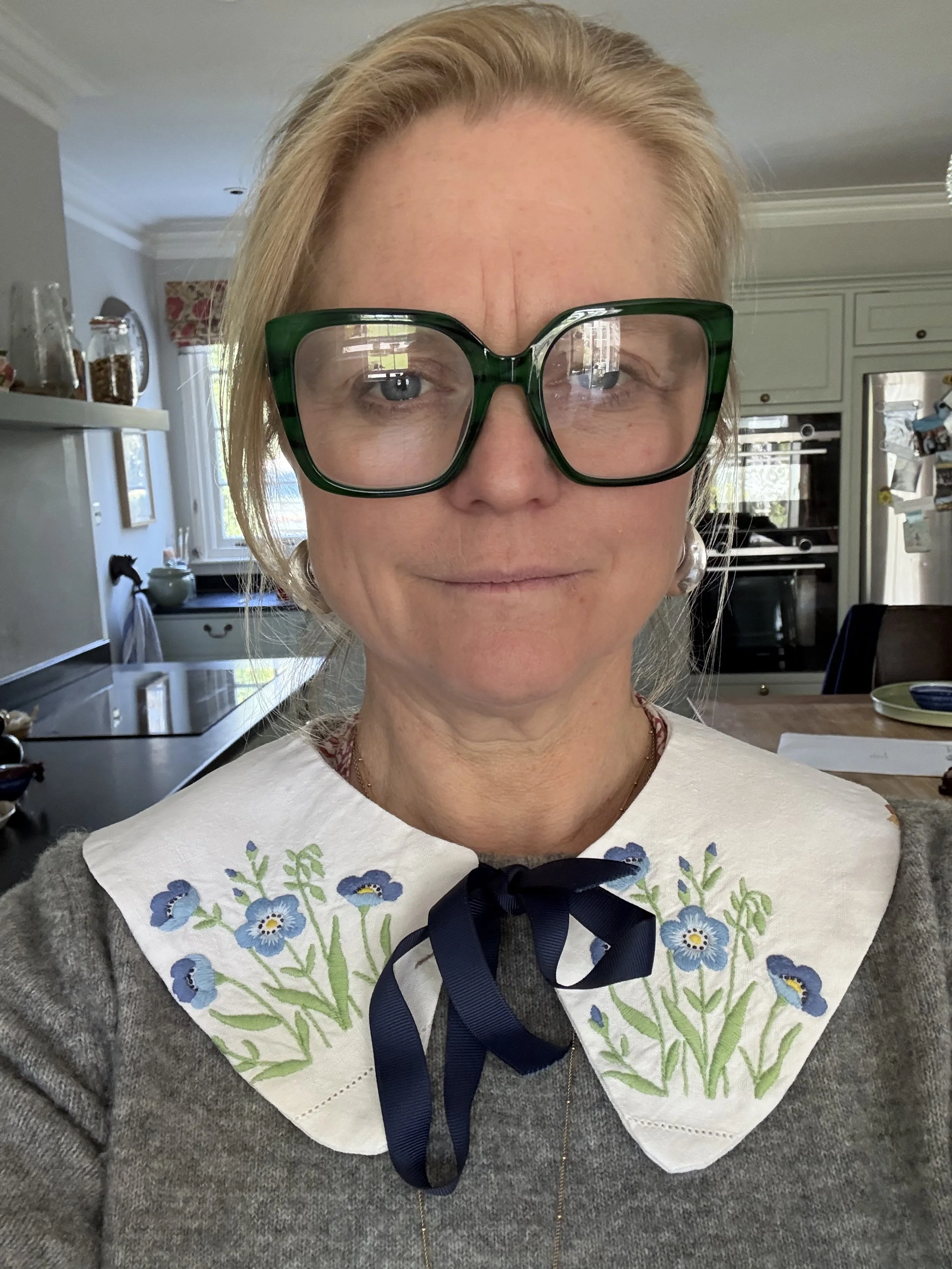 A woman with blonde hair, large green glasses, and wearing a embroidered white collar with blue flowers and a blue bow, standing in a kitchen interior.