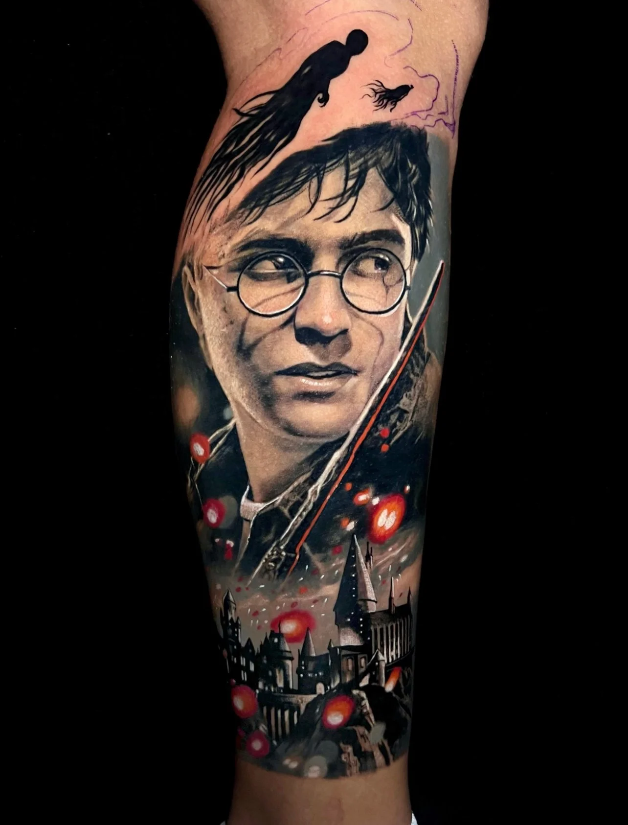 Tattoo of a young man's face with glasses, dark hair, and a serious expression, set against a dark cityscape with spires and glowing red orbs, and a silhouette of a figure with a wand or staff at the top.