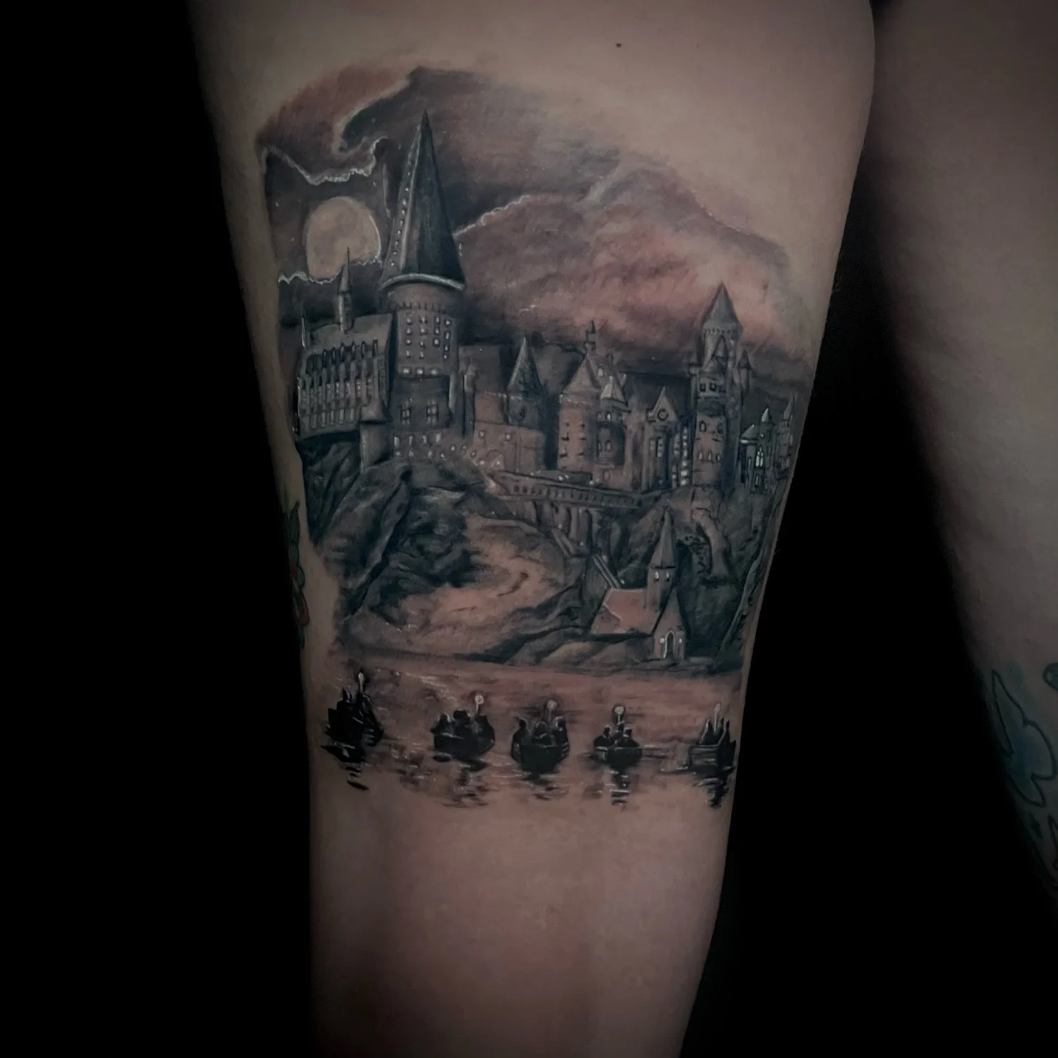 Black and gray tattoo of a castle with multiple towers and turrets, set on a hill overlooking a river with boats and figures, under a cloudy sky with a full moon.