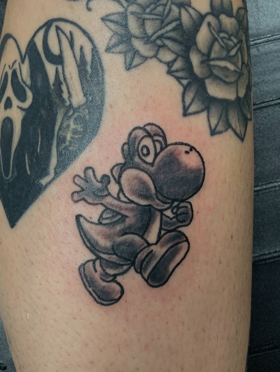 A black and gray tattoo of Yoshi, a character from Super Mario, on a person's arm, with tattoos of roses and a skull and crossbones.