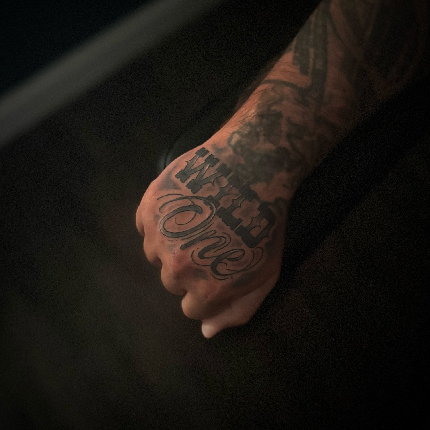 Close-up of a person's hand with a tattooed arm, the tattoo on the hand reads 'Family' in a decorative script, with additional tattoos visible on the arm.