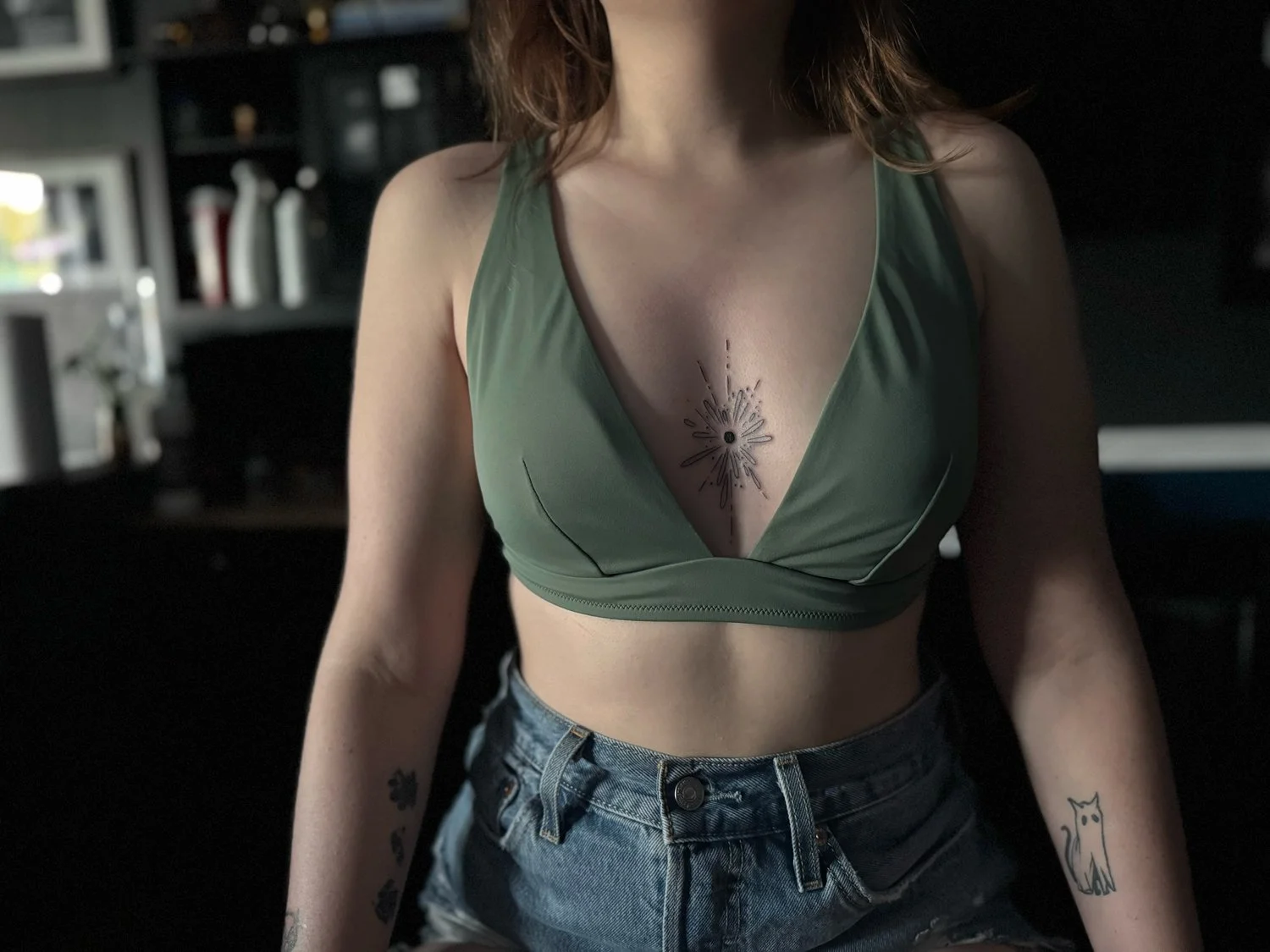 A woman with tattoos on her arms and chest wearing a green sports bra and denim shorts, standing in a dimly lit room.