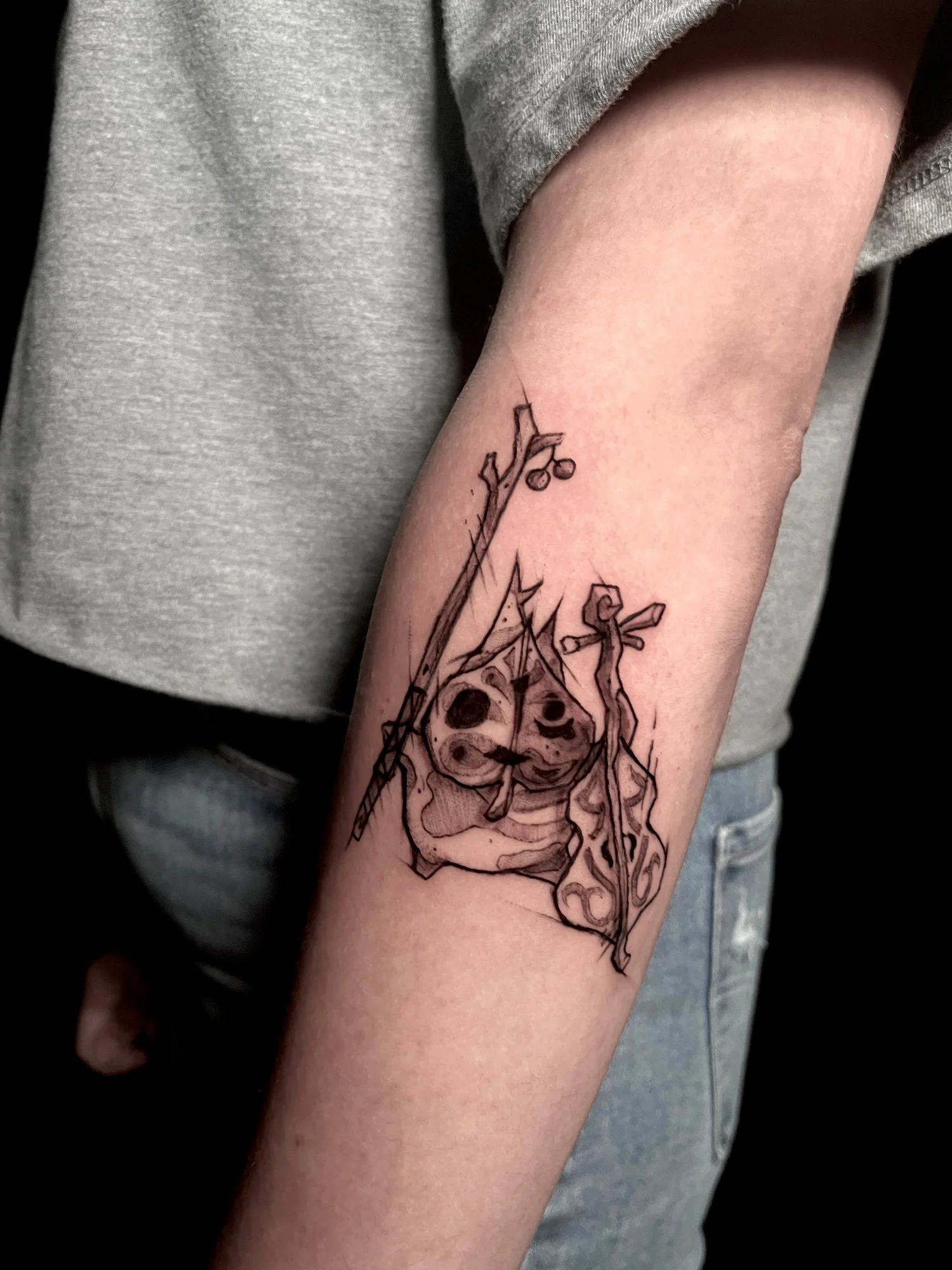 Black ink tattoo of a hanging skull, a crossed spoon and fork, and a textile that looks like a cloth or flag on a person's forearm.
