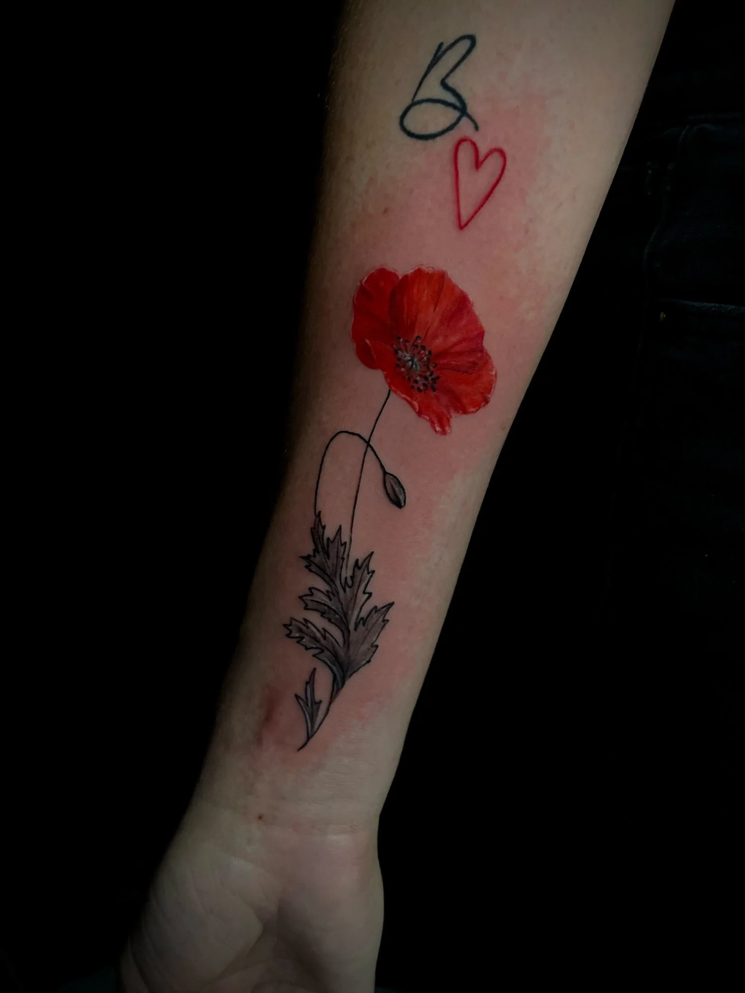 Tattoo of a red poppy flower with black details on the petals, along with a black and red outline of the word 'B' and a heart symbol above the flower.