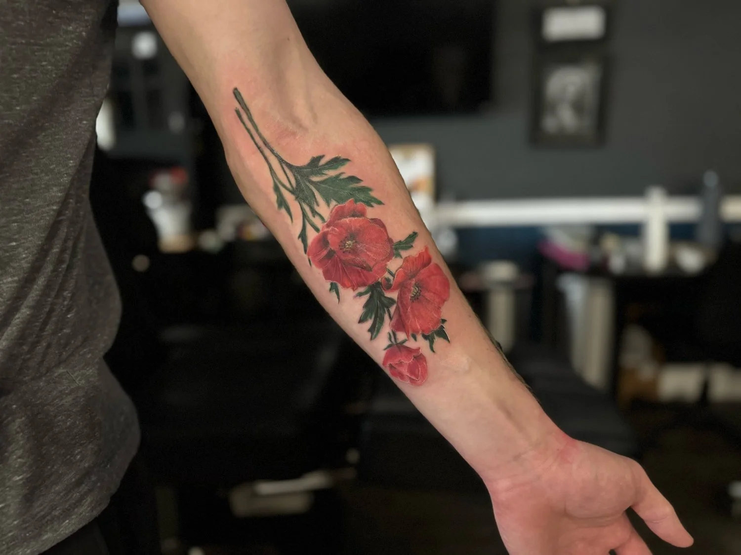 Tattoo of red flowers and green leaves on person's forearm inside a room.