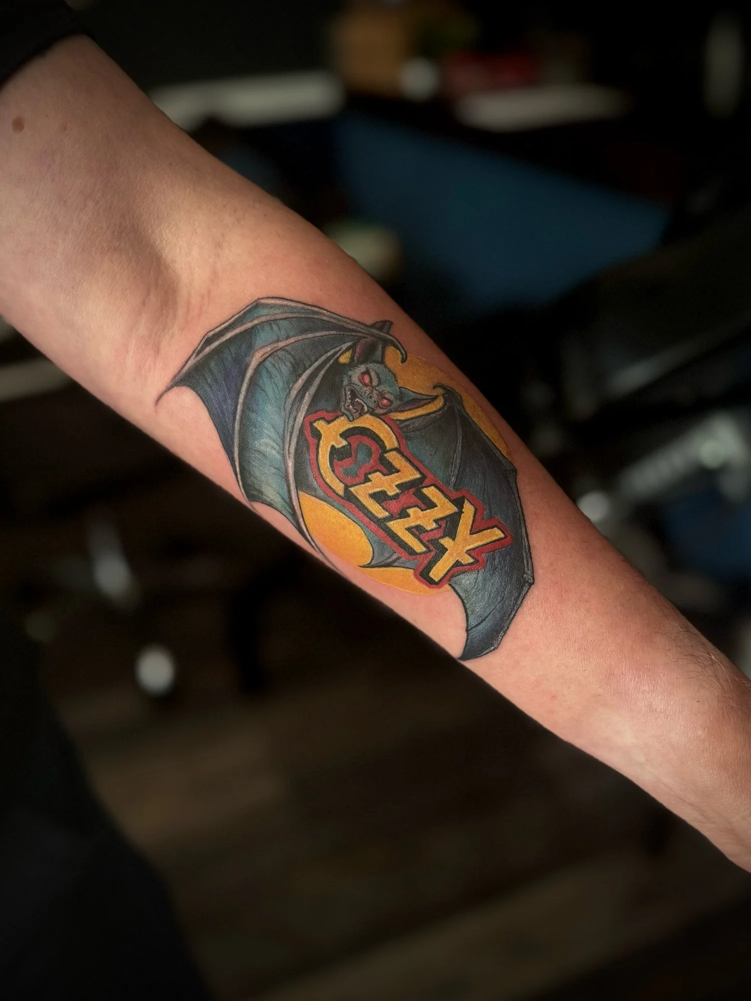 Tattoo of a bat with red eyes and spread wings, holding a yellow moon with the word 'XIZ' in red and yellow letters.