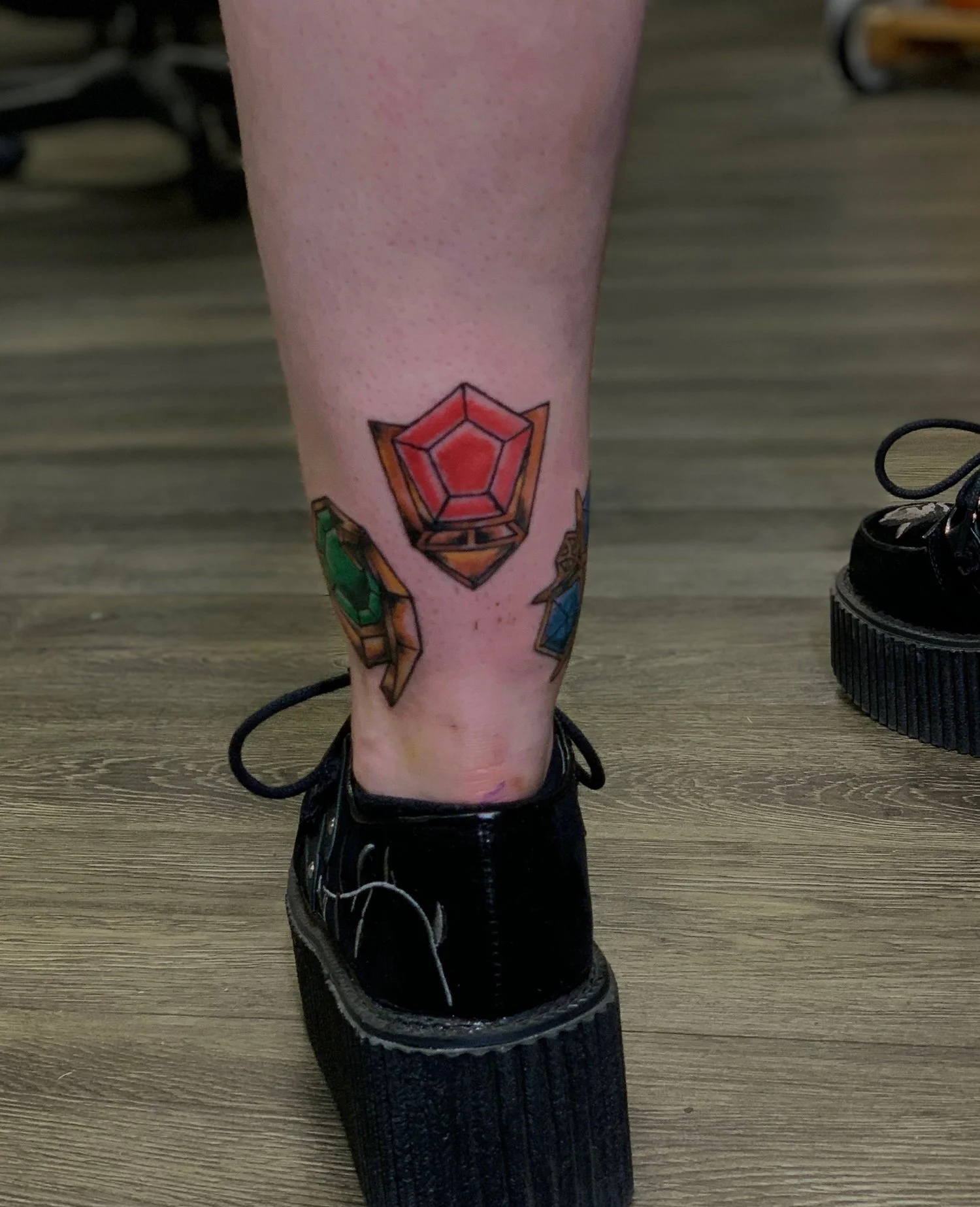 Tattoo of a pink and red gemstone on a person's lower leg, surrounded by smaller colorful gemstones.