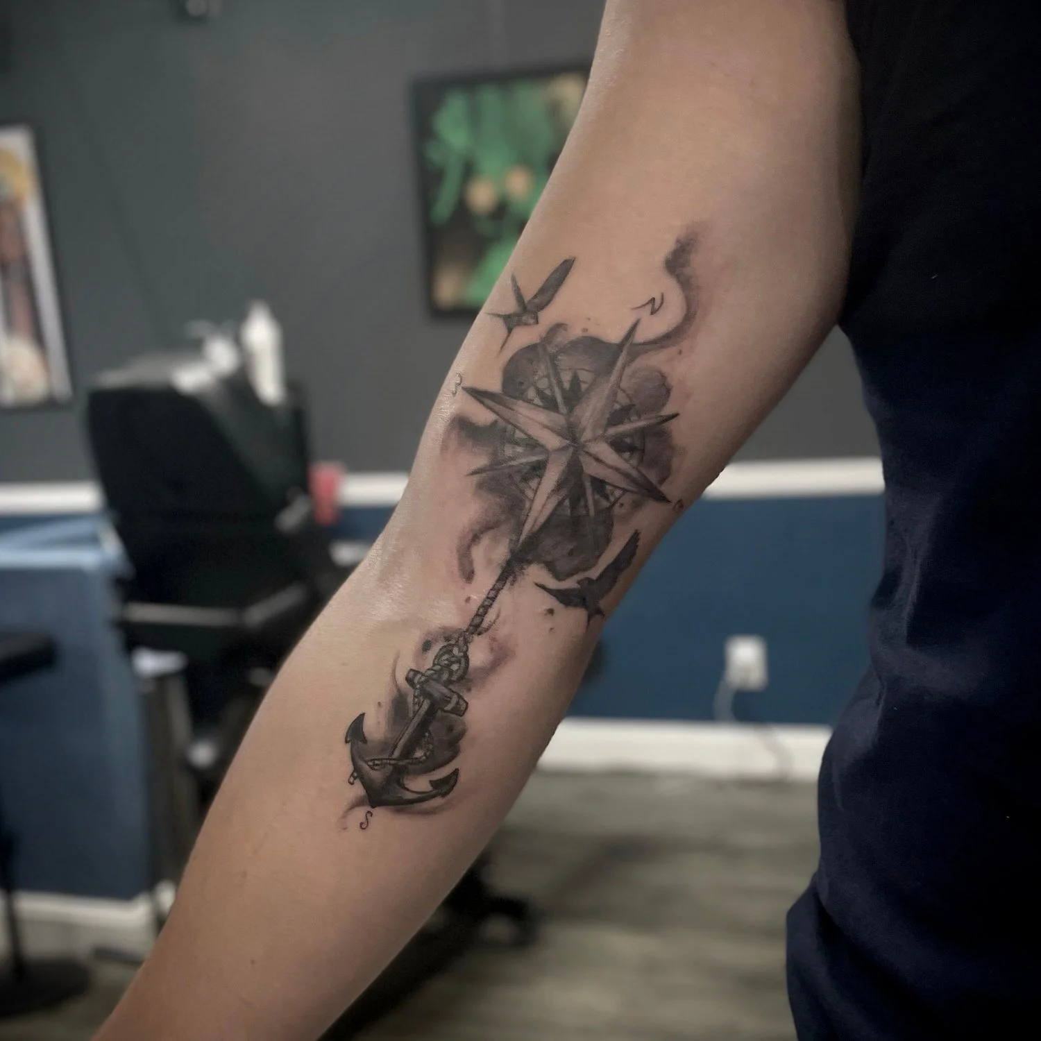 Black and gray tattoo of a compass with an anchor hanging from it on the person's forearm in a tattoo studio.