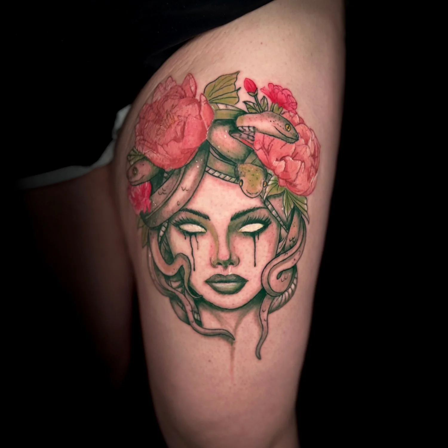 Tattoo of a woman's face with closed eyes, tears, and snakes in her hair, surrounded by pink flowers and green leaves.