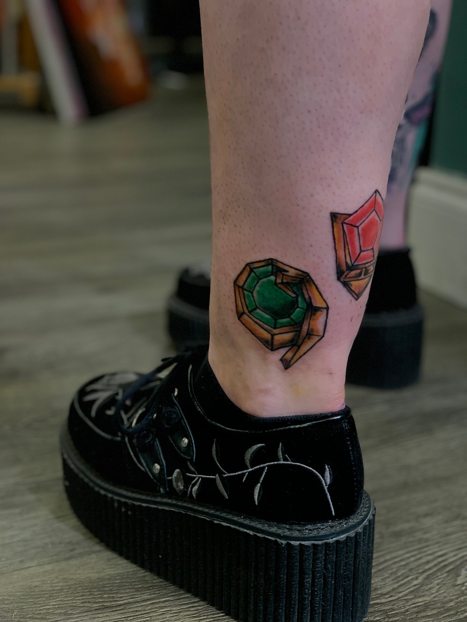 Colorful tattoo of a gemstone and a ring on person's ankle, with black platform shoes and a wooden floor.