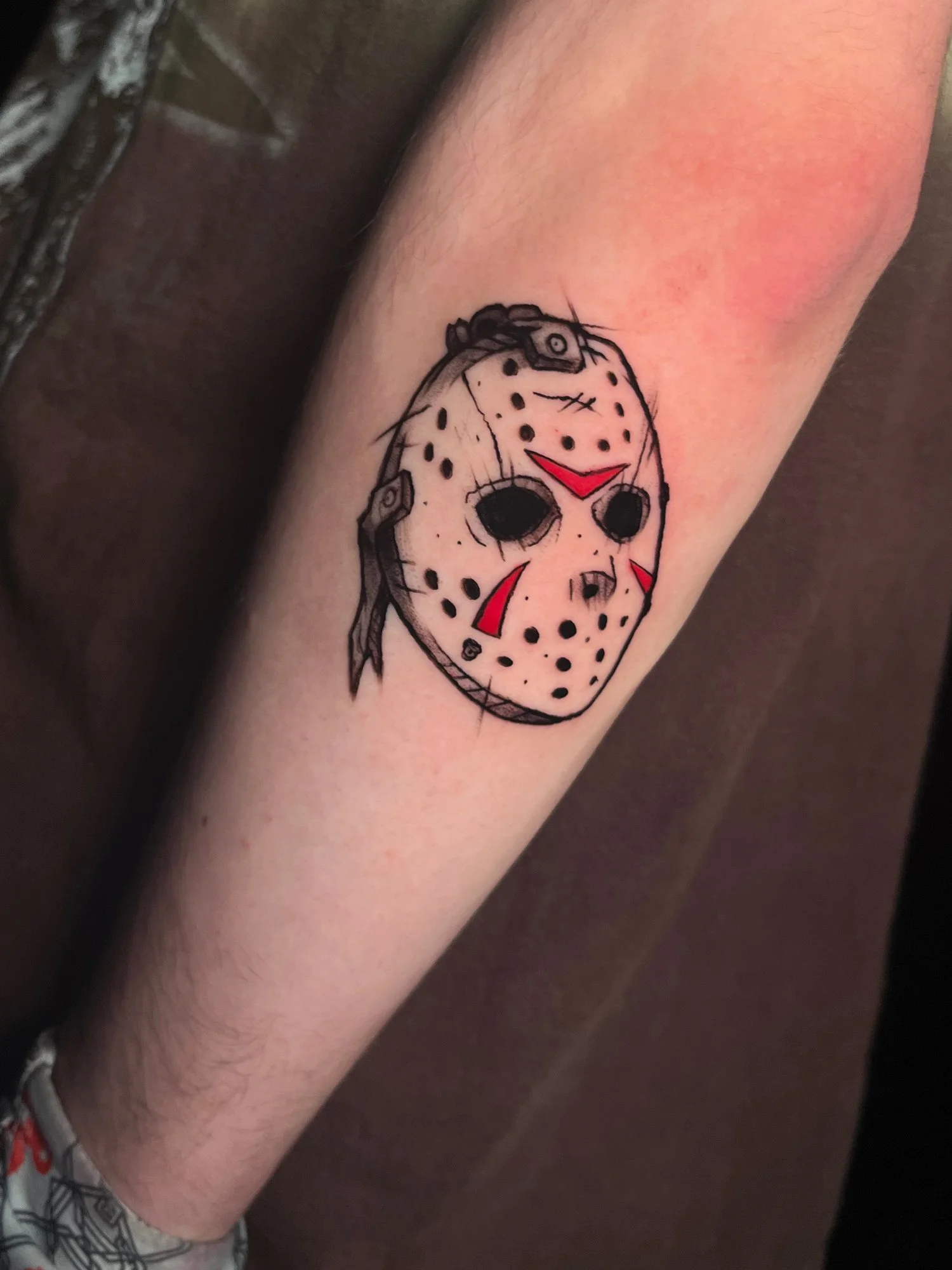 Tattoo of a cartoonish hockey mask with black holes for eyes and red accents on the face, resembling Jason Voorhees from horror movies.