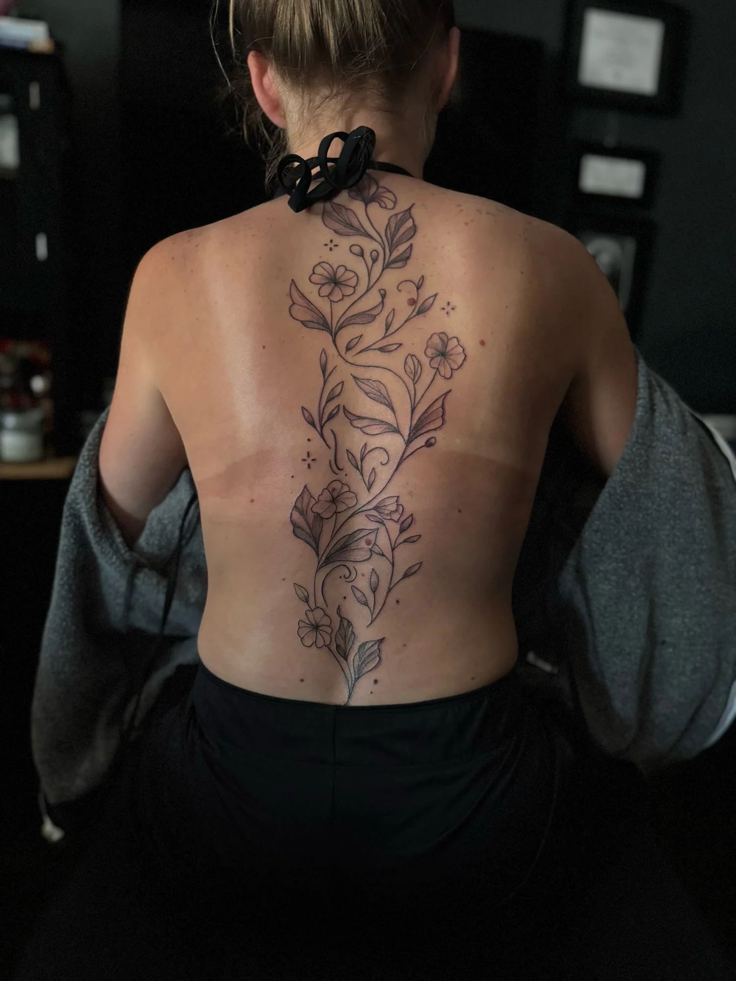 Back view of a woman with a long floral tattoo running from her upper back to lower back, featuring flowers, leaves, and swirling vines.