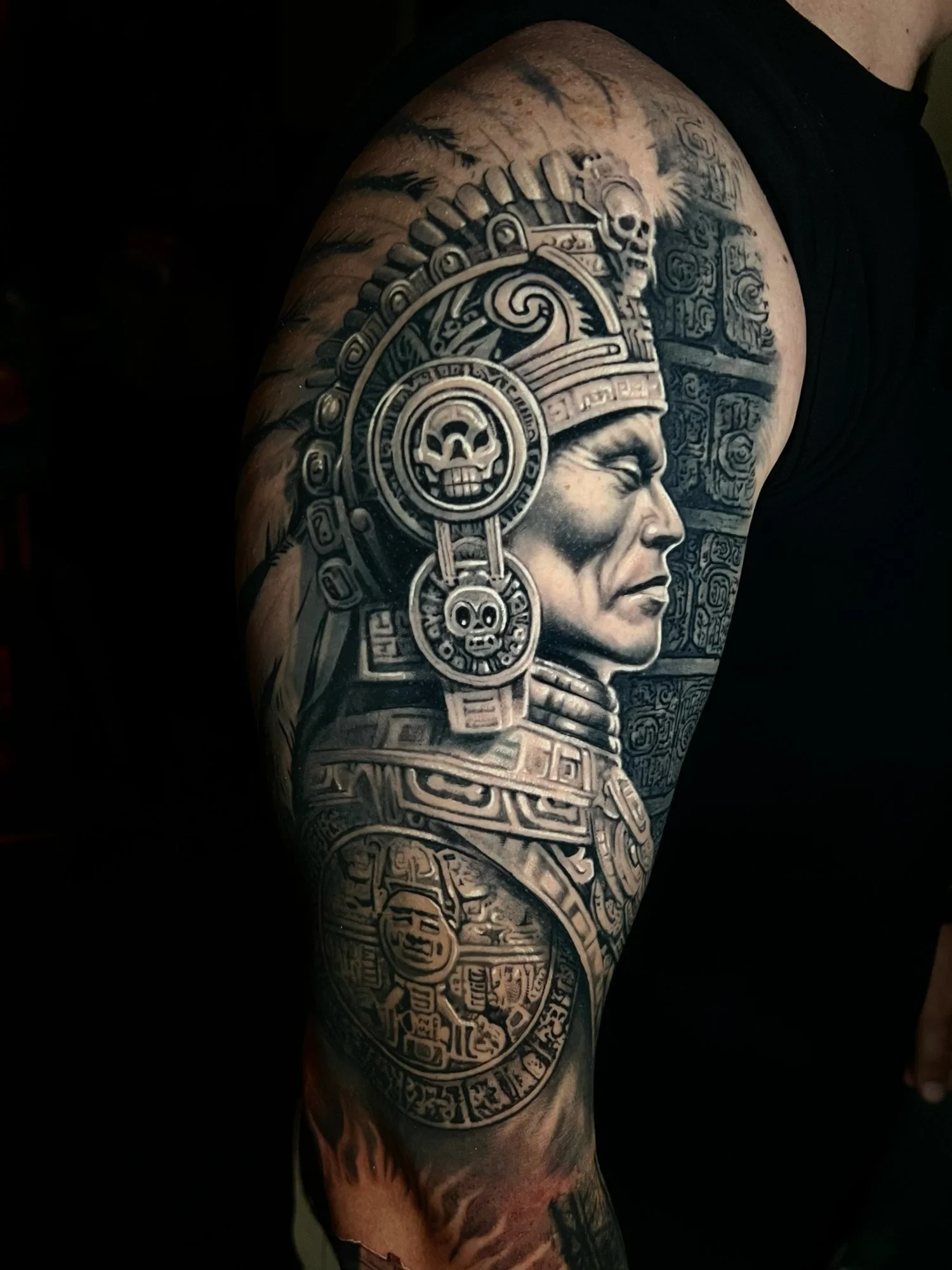 Black and gray tattoo of an Aztec figure with a headdress featuring skulls, along with Aztec symbols and inscriptions, covering the upper arm.