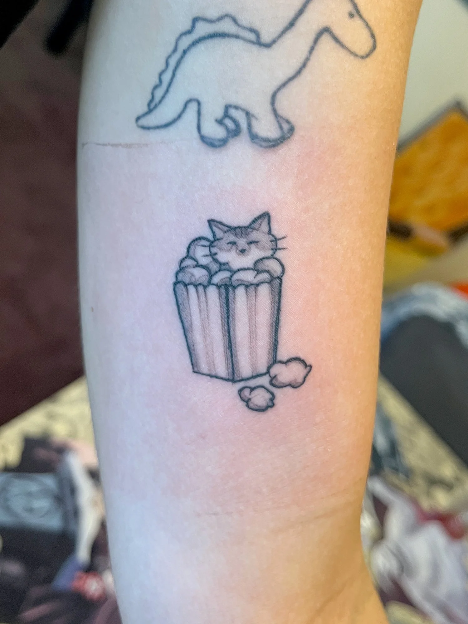 Tattoo of a cartoon cat sitting inside a cupcake with sprinkles, with a small puff of smoke or steam at the bottom.