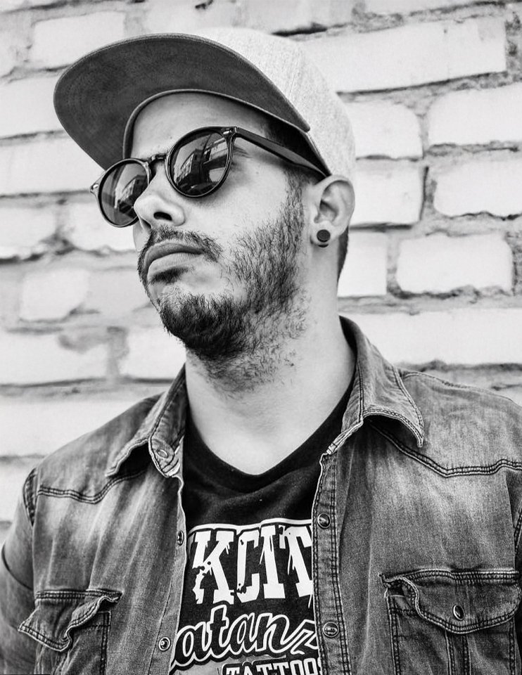 Osmel Rodriguez Black and white photo of a young man wearing sunglasses, a baseball cap, a denim jacket, and a graphic T-shirt, standing in front of a brick wall.