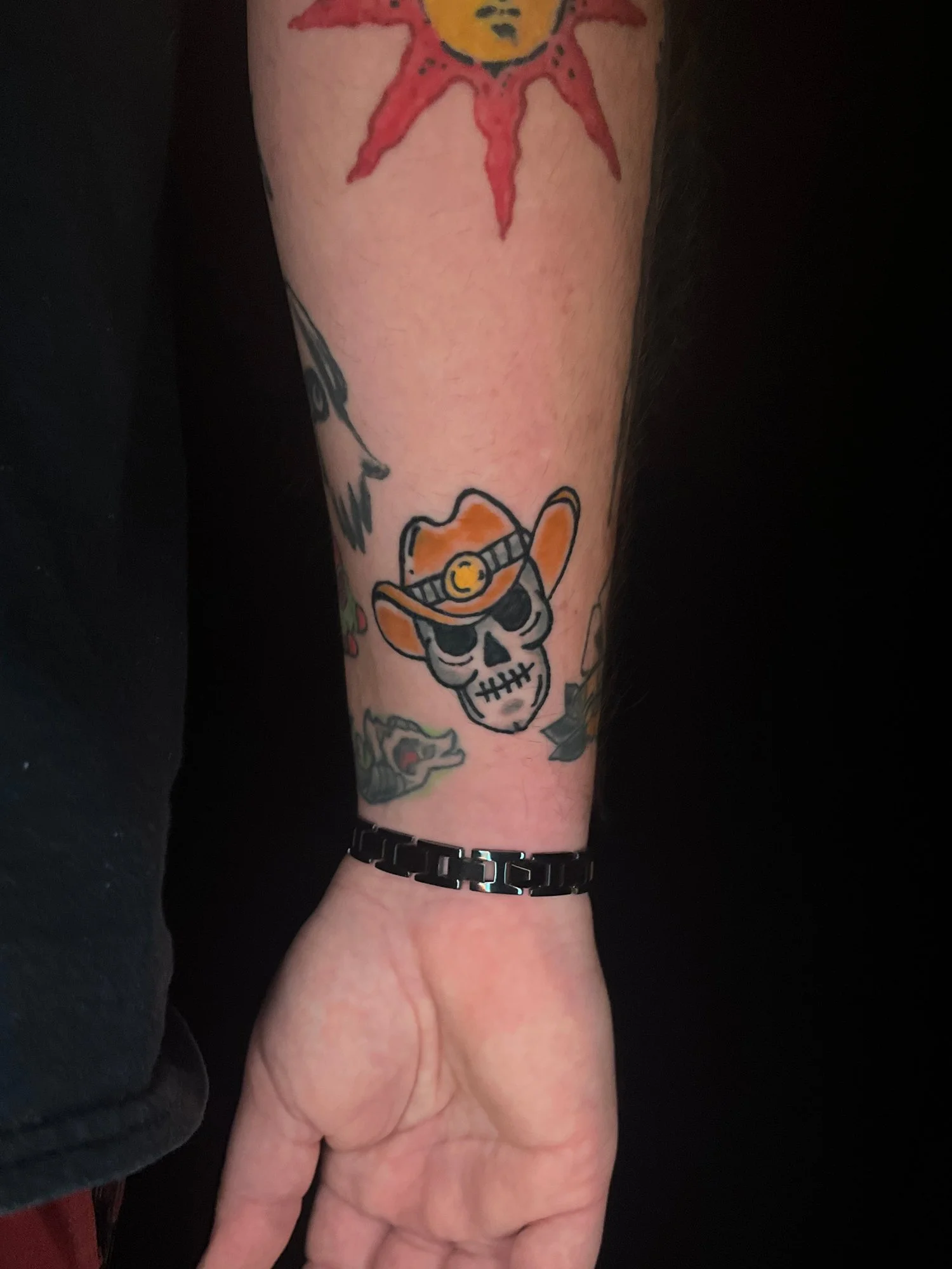 Tattoo of a skull wearing an orange cowboy hat and a headlamp on a person's forearm, with additional tattoos partially visible around it.