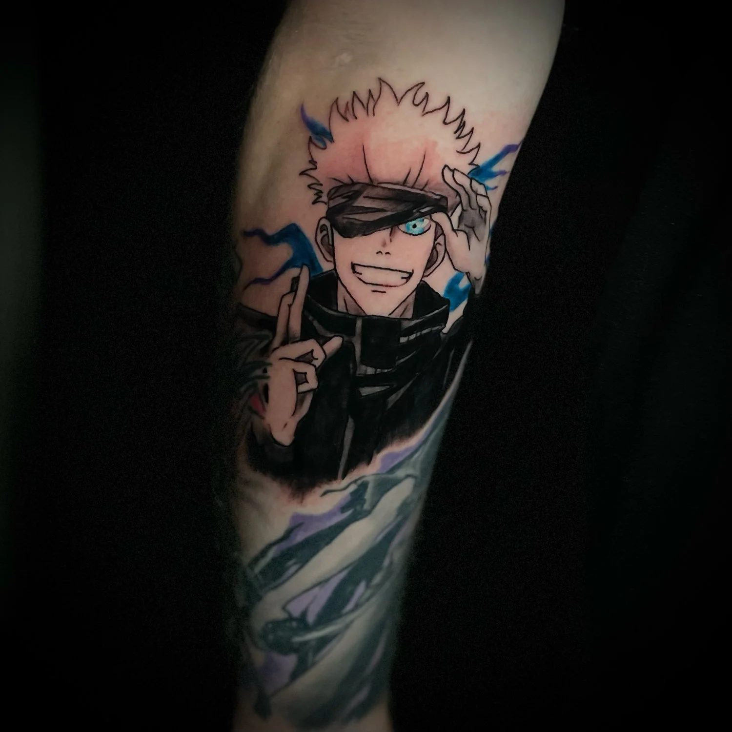 Tattoo of a comic-style character with light hair, blue eyes, wearing black clothing, and covering one eye with blackout glasses. The character is smiling and making a gesture with their fingers. Blue energy or lightning effects surround the characte