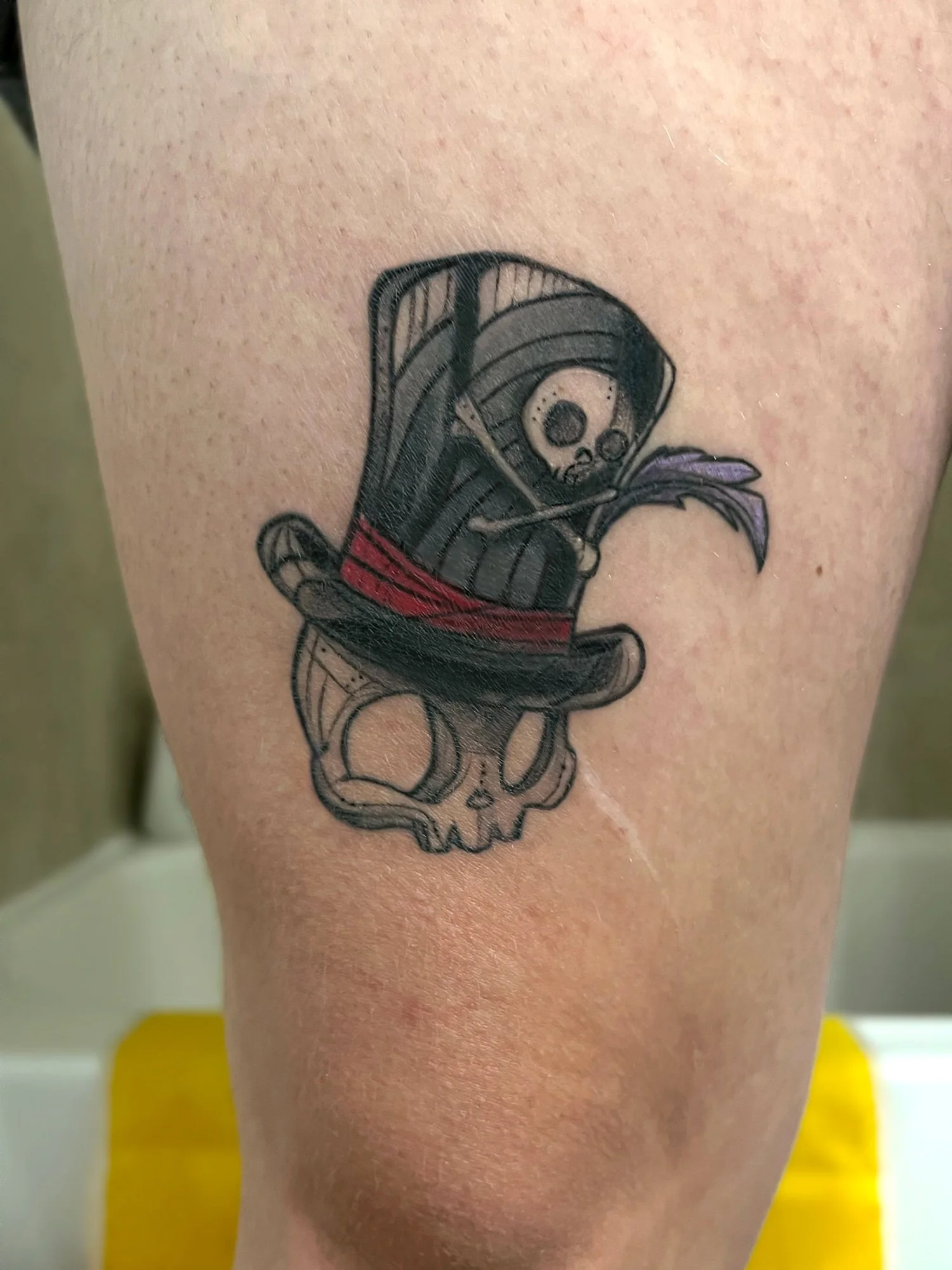 Tattoo of a skeleton with a skull holding a feather and sword, wearing a black hat with a red band, above a skull outline.
