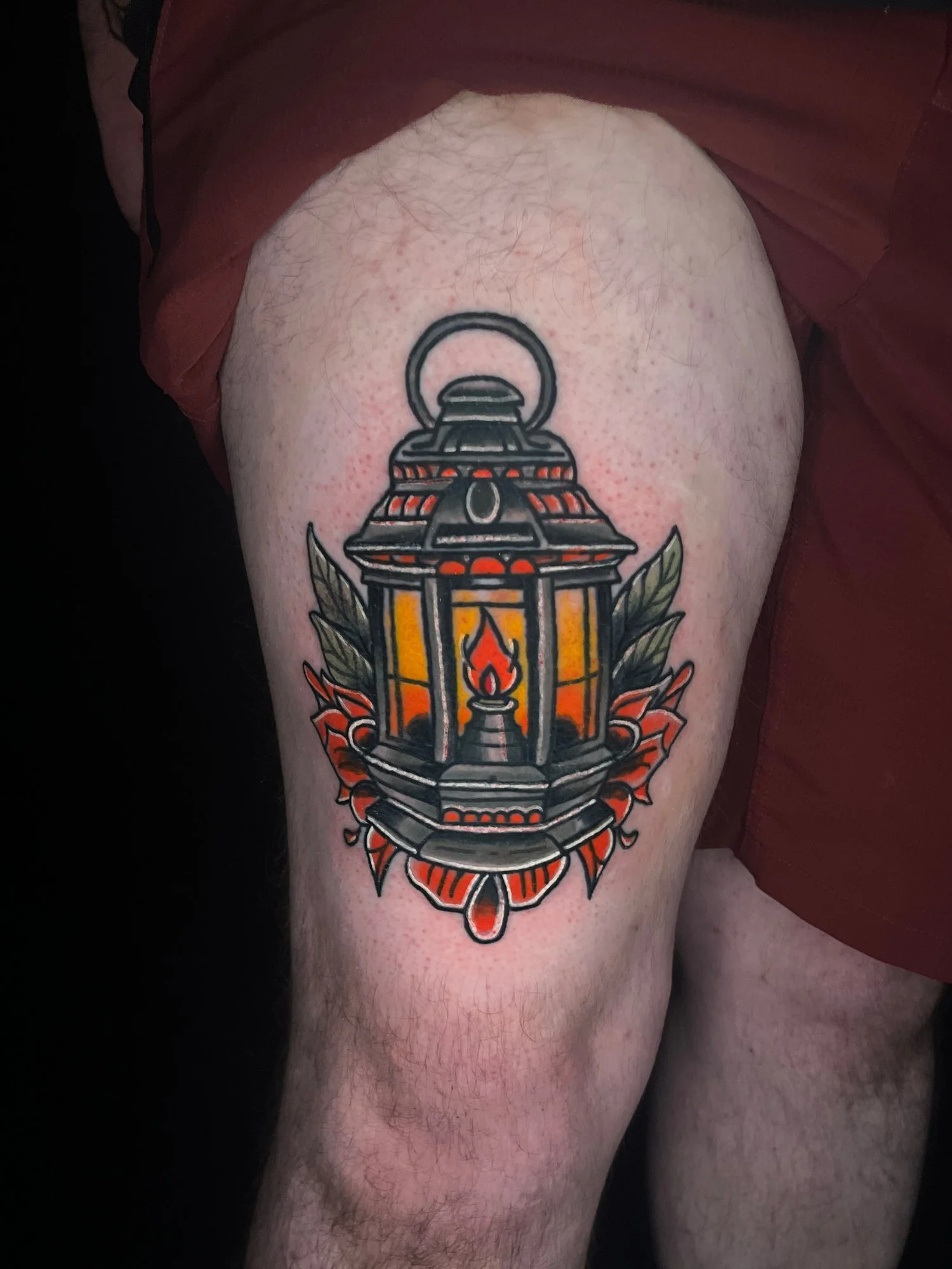 Colorful tattoo of a lantern with a flame inside, surrounded by leaves and decorative elements, on a person's thigh.