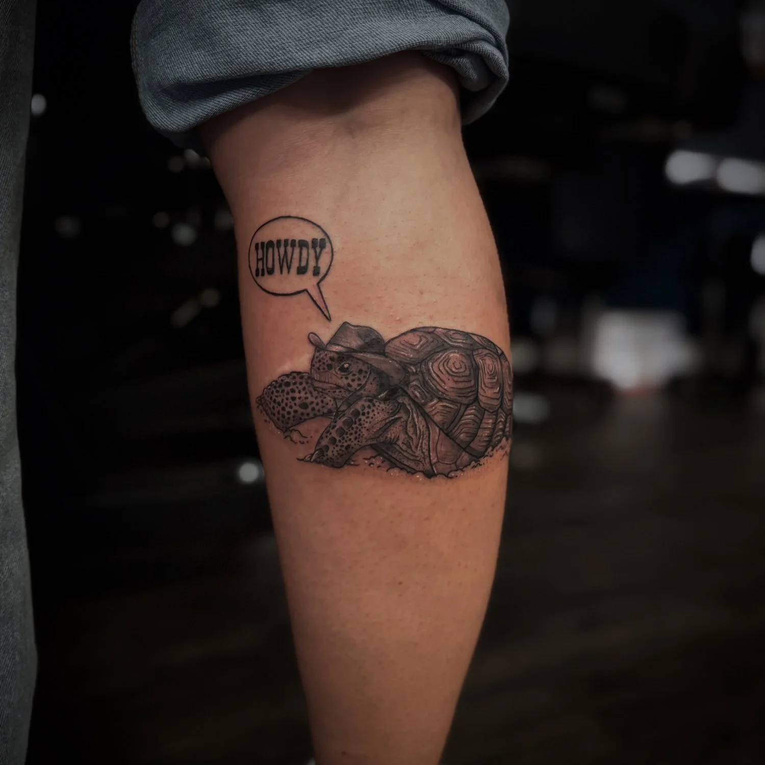 Tattoo of a turtle with a speech bubble saying "HOWDY!" on a person's arm.
