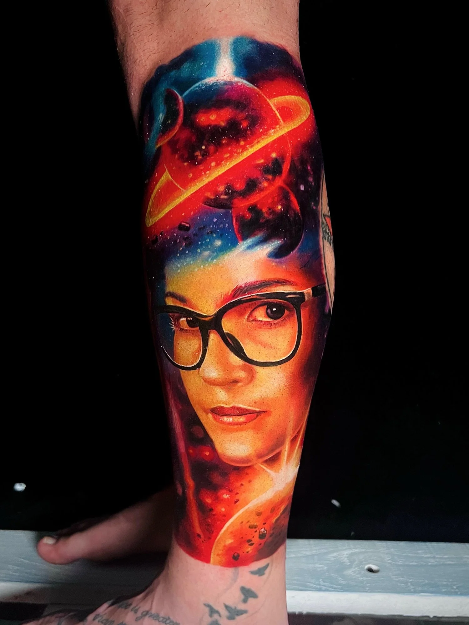 Colorful tattoo of a woman's face with glasses, space scene with planets and stars, and abstract swirling patterns.