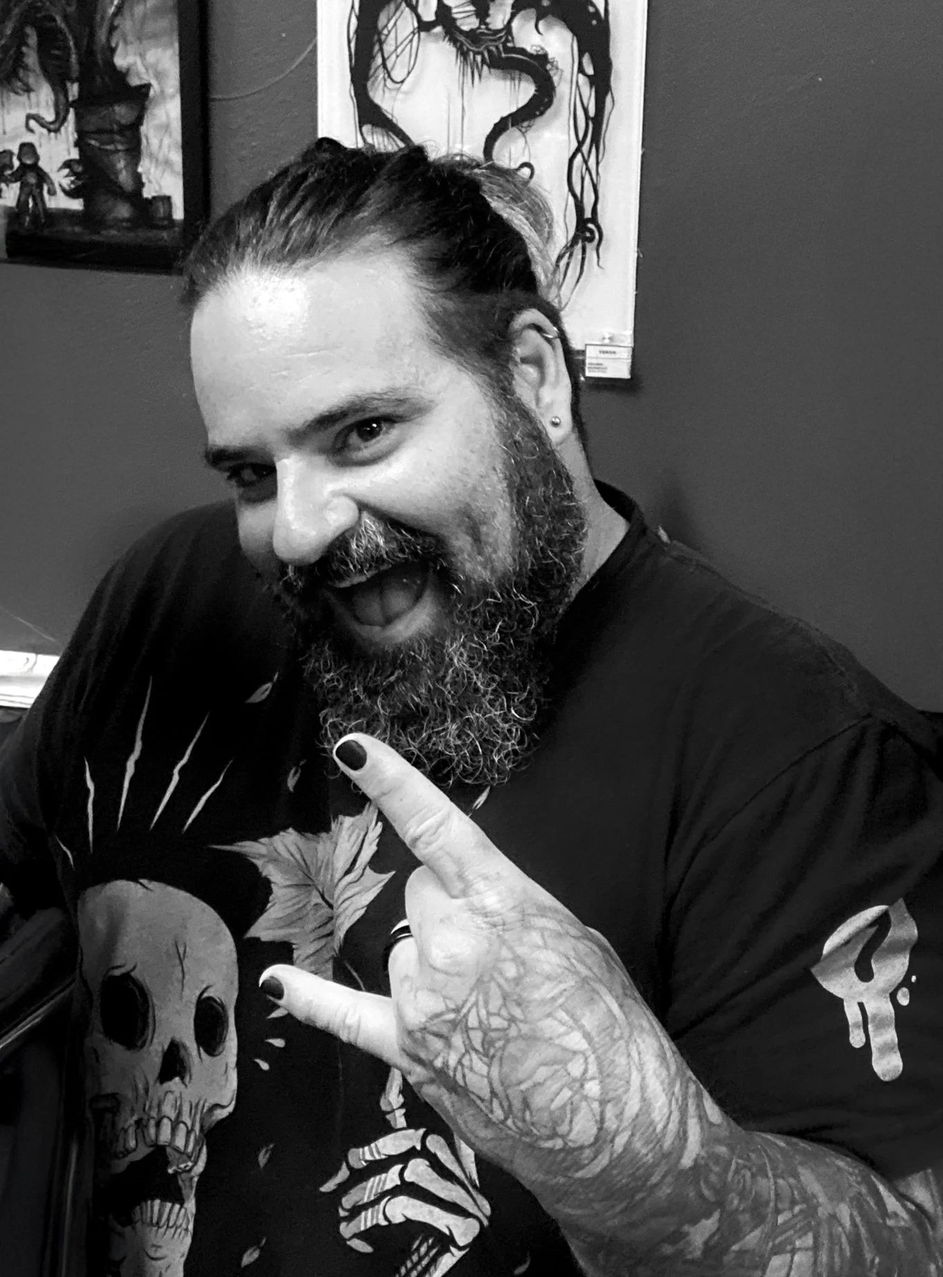 Will Pigg with a beard and long hair tied back, smiling and making a rock hand gesture. He has tattoos on his arm and is wearing a black shirt with skull and skull illustration designs. Background contains framed artwork on the wall.