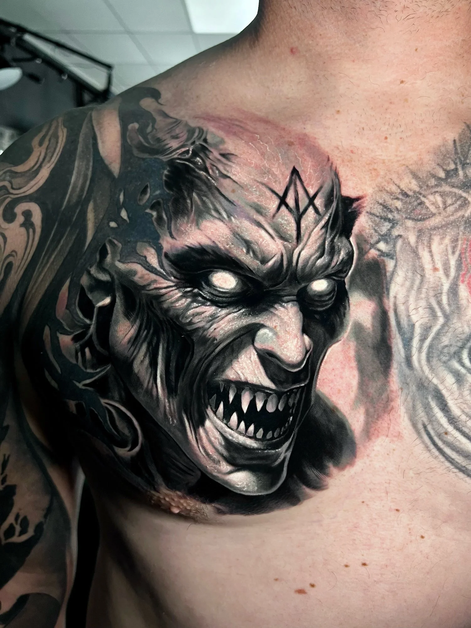 Close-up of a person's upper chest tattoo depicting a fierce, monstrous face with sharp teeth, glowing eyes, and dark shading.