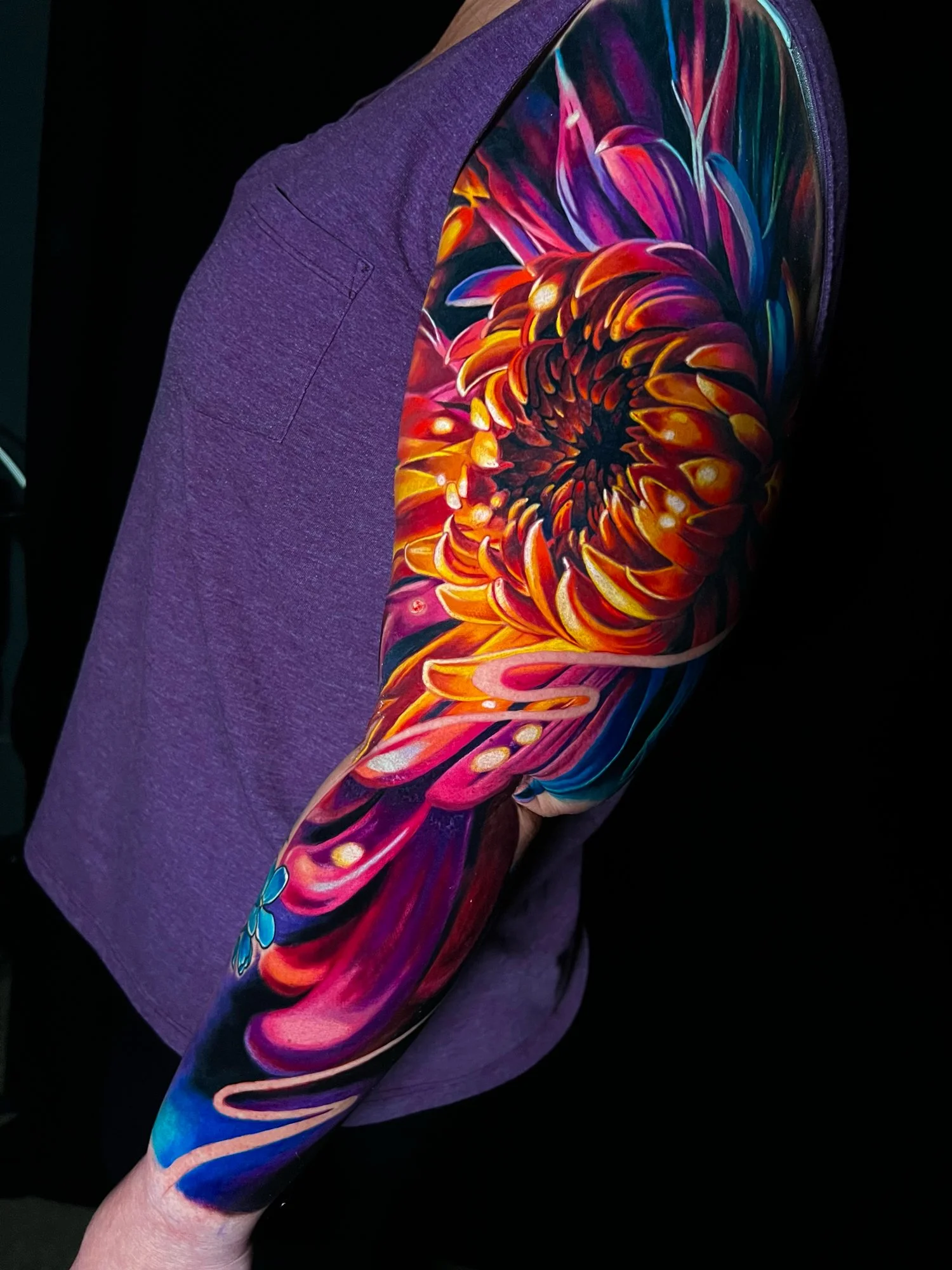 A colorful, vibrant tattoo of a floral design on a person's upper arm and shoulder, featuring swirling patterns in shades of red, orange, yellow, pink, purple, and blue.