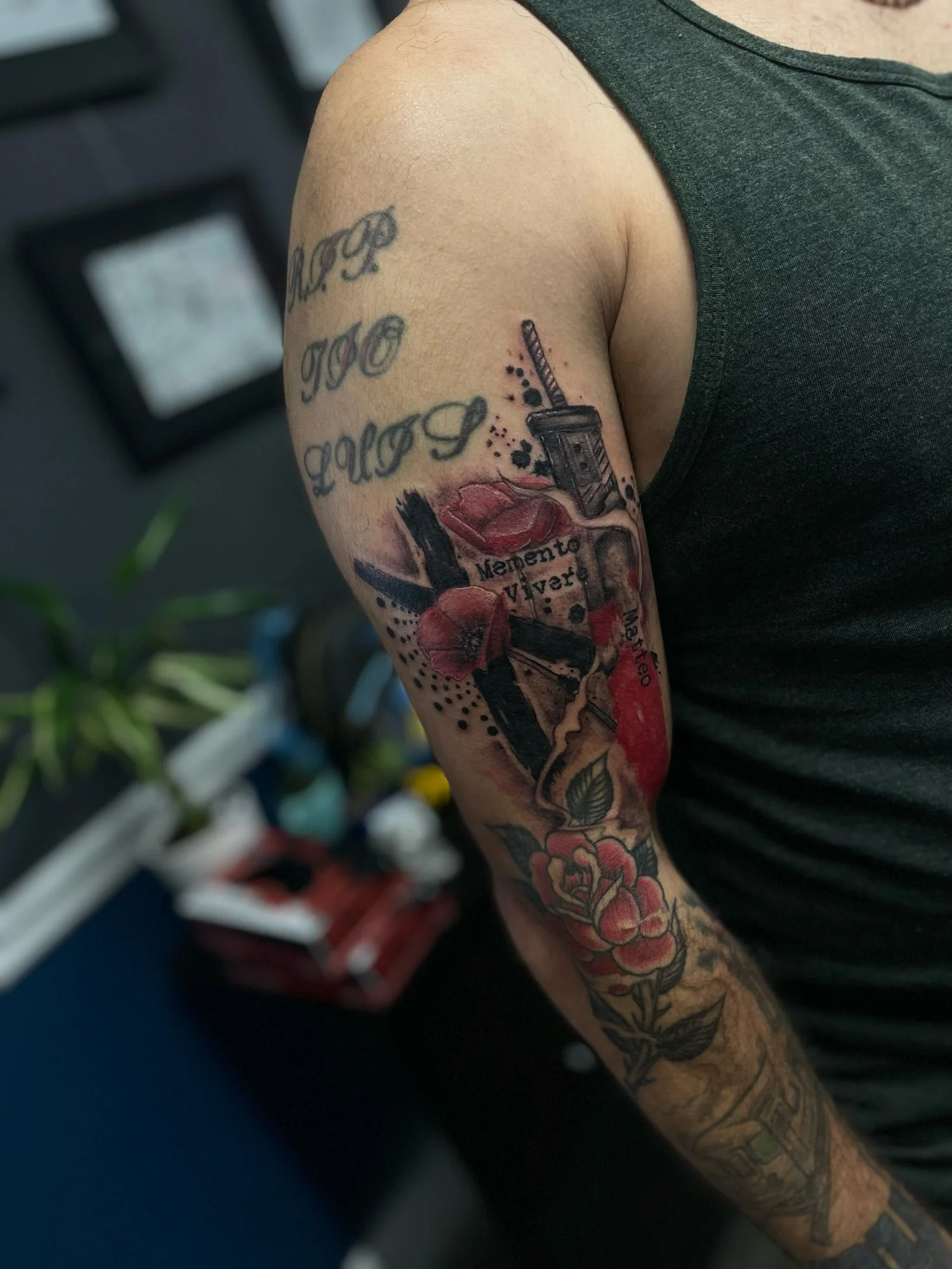 Tattooed upper arm with a black tank top, featuring a tattoo of a red flower, lipstick, and makeup items, with a background of blurred office or studio setting.