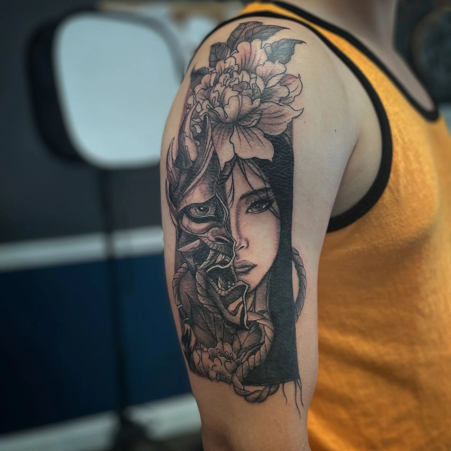 Tattoo of a woman's face with floral elements and mythical creature, in black and gray ink on upper arm.