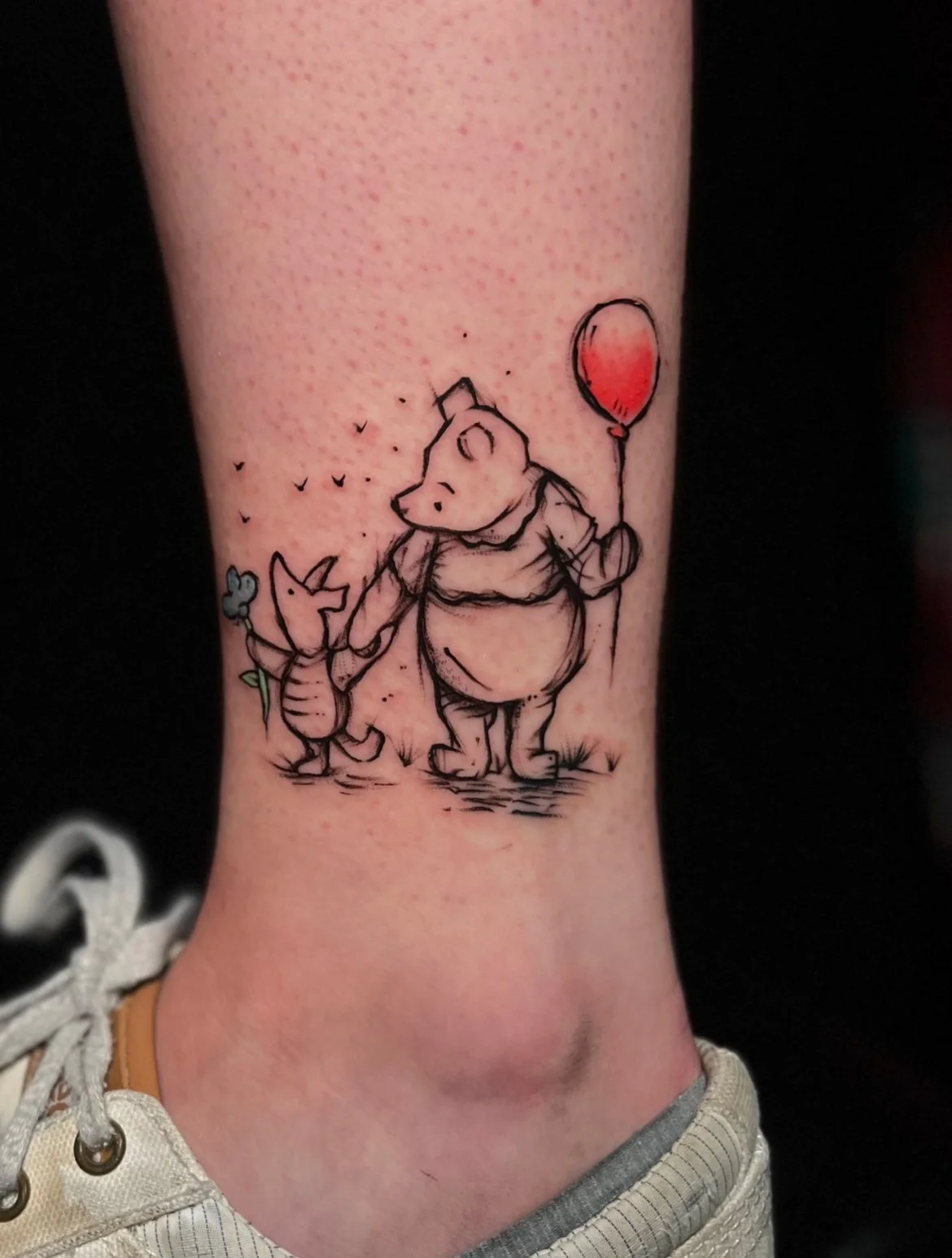 Tattoo of Winnie the Pooh and Piglet holding hands, with Piglet holding a red balloon, on a person's ankle.