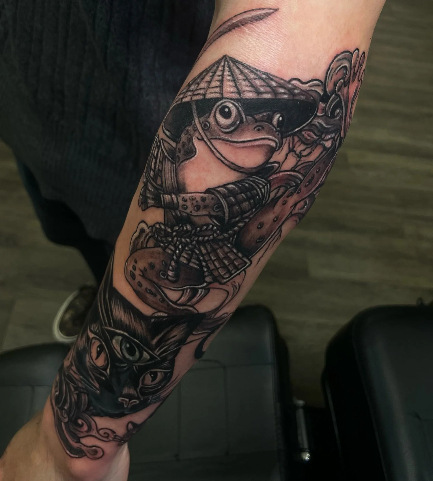A black and gray tattoo of a frog with a bamboo hat and sword, and a cat with a third eye, on a person's forearm.
