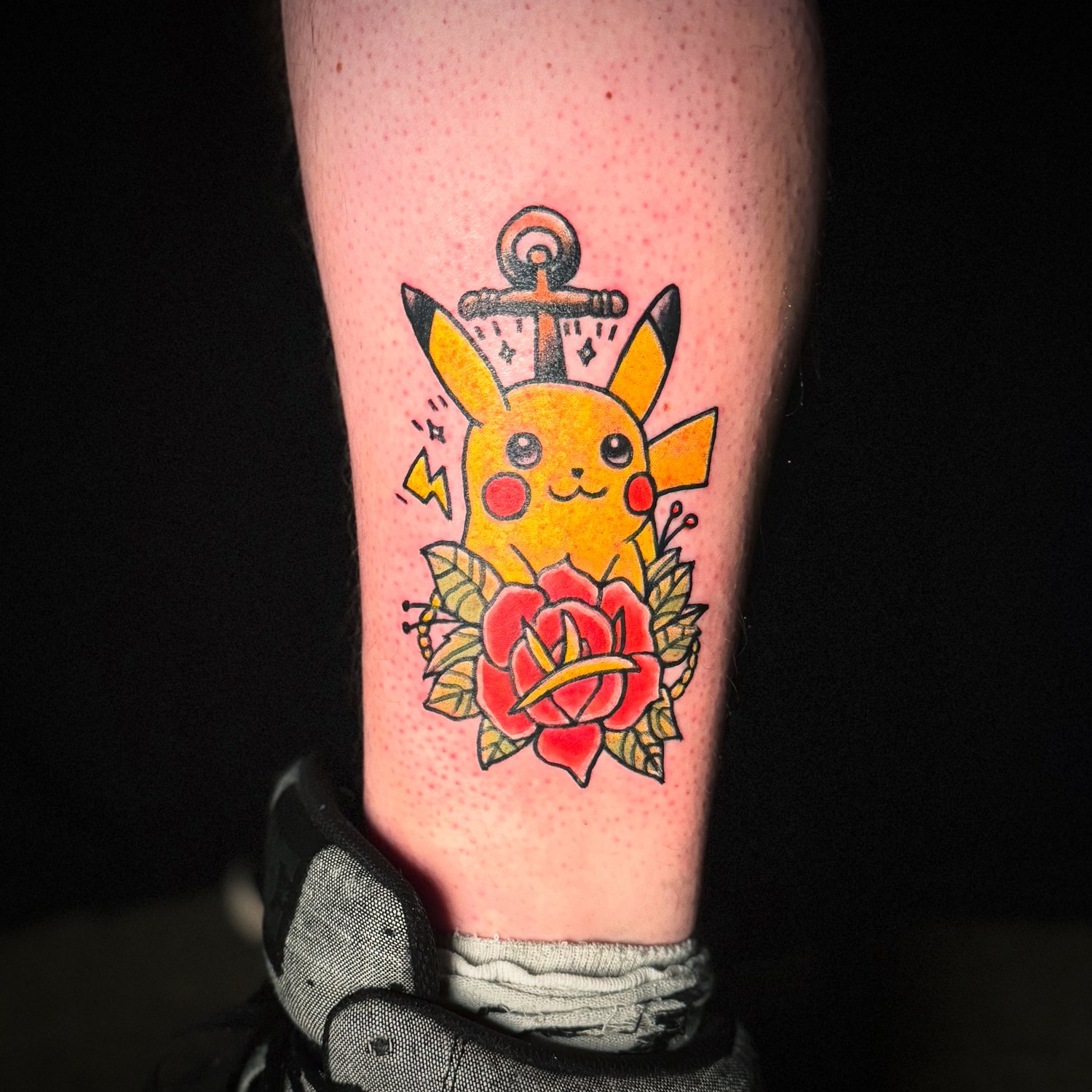 Tattoo of Pikachu with a sword behind its head, a lightning bolt, surrounded by red roses and green leaves.