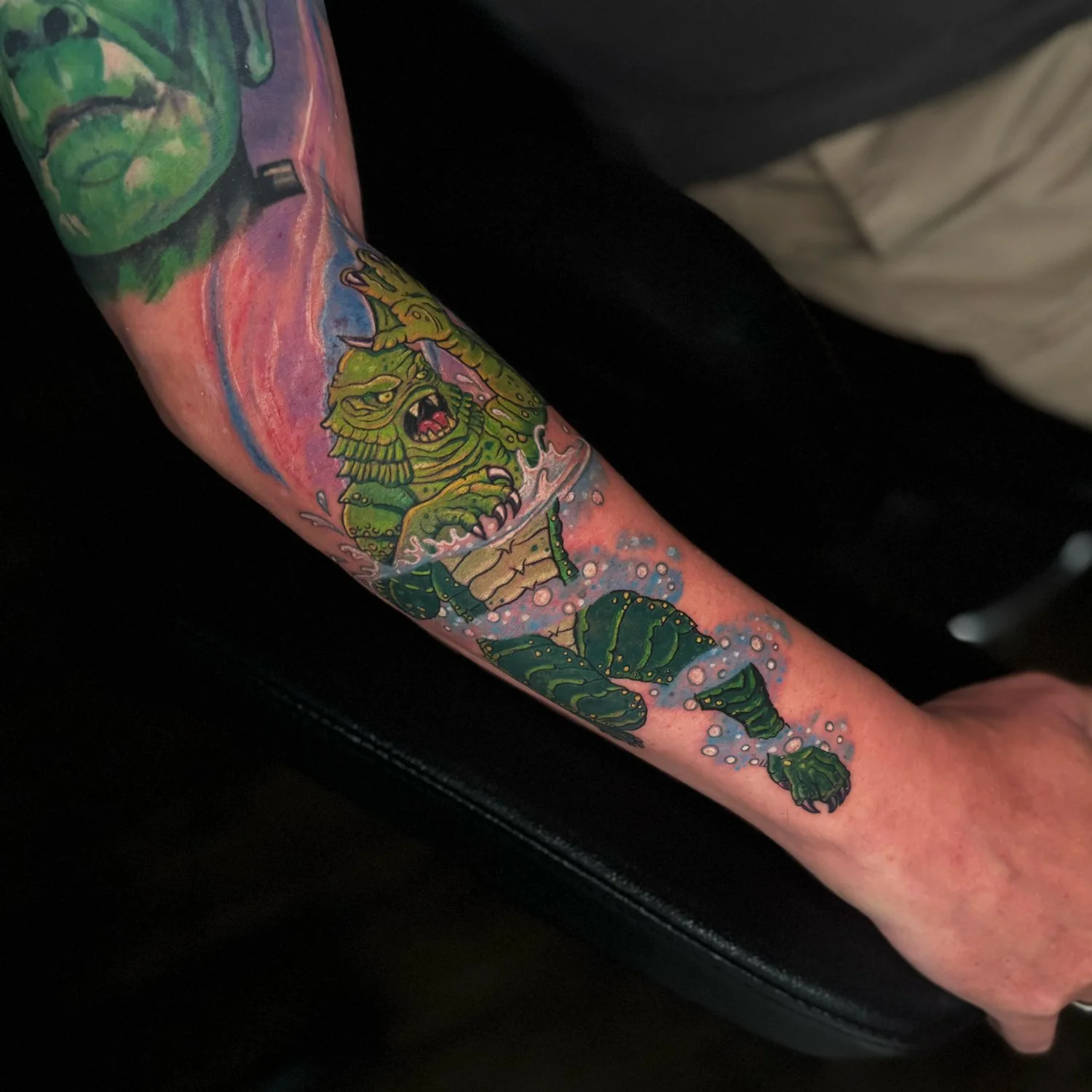 Colorful tattoo on forearm depicting a fierce green monster emerging from water, with bubbles and aquatic plants.