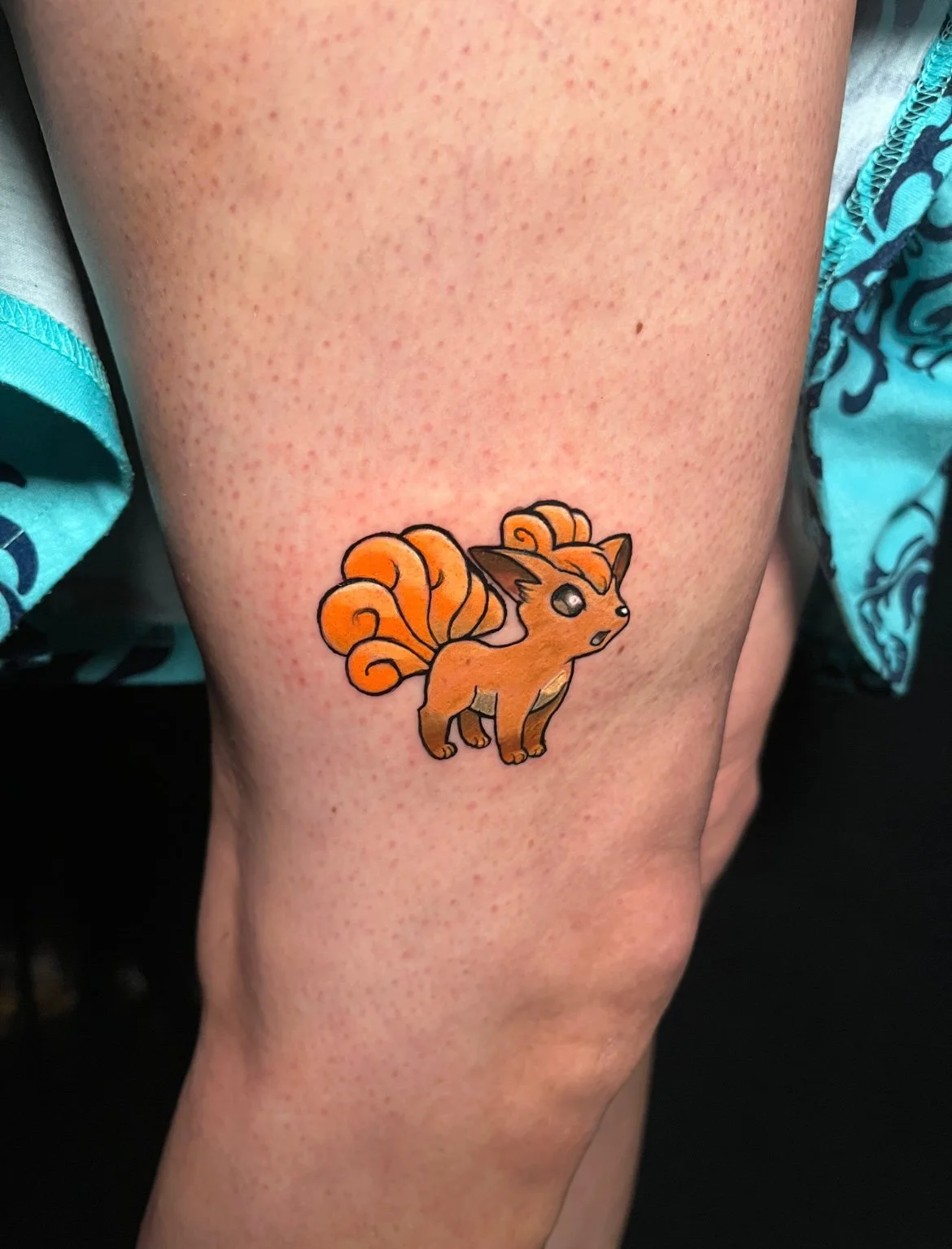 Close-up of a person's thigh with a colorful fox tattoo featuring orange fur, a curled fluffy tail, pointed ears, and wide eyes.