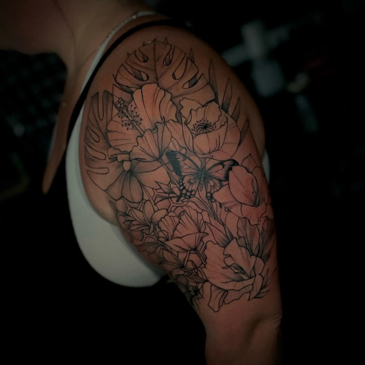 A shoulder tattoo featuring outlined flowers, a butterfly, and Monstera leaves.