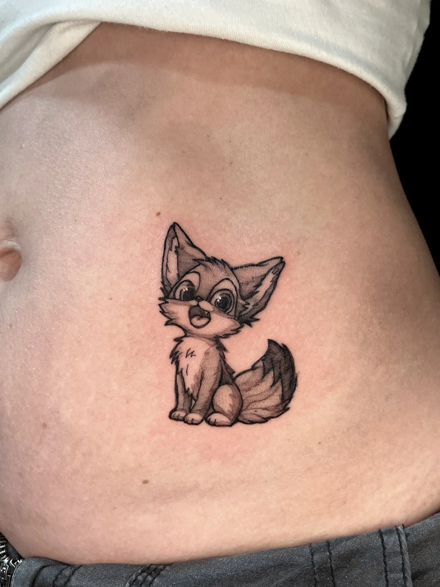 Tattoo of a cute cartoon fox sitting on a person's side, looking surprised or excited.