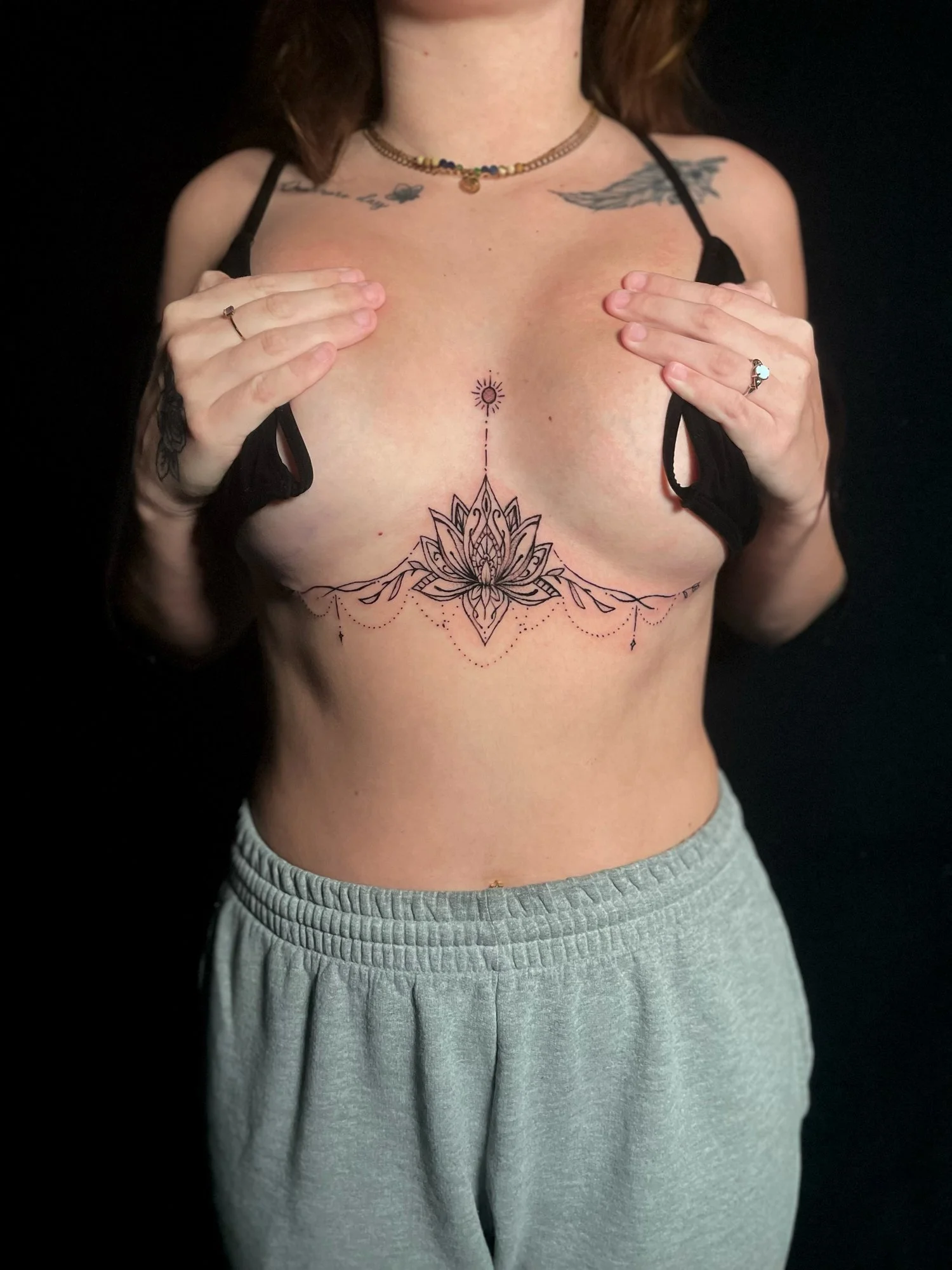 Woman with tattoos holding her breasts, showing a large lotus flower tattoo across her chest, with decorative dots and lines, against a black background.